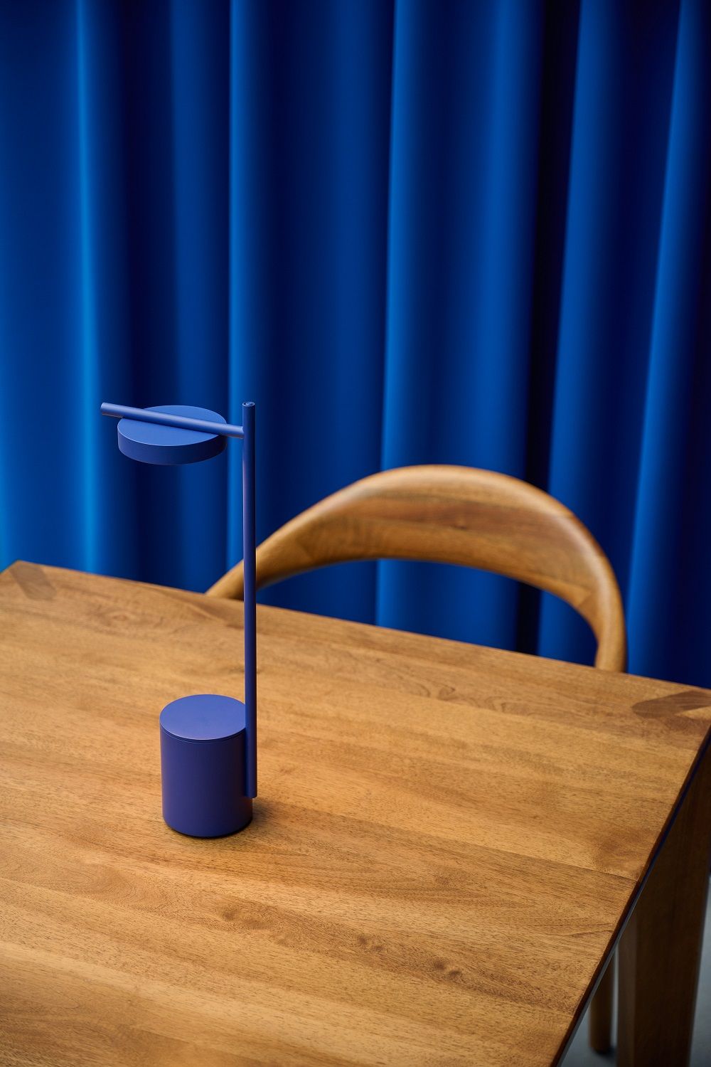 Blaue Igram Akku-Tischleuchte von Grupa auf Holztisch vor blauem Vorhang und Stuhl.