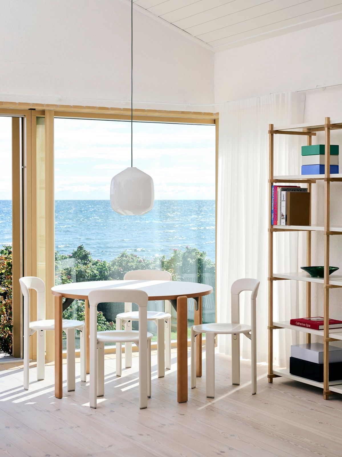 Helle Esszimmerszene mit Hay Pendelleuchte Aspekt Buoy Glass, Esstisch und Stühlen vor großem Fenster mit Meerblick.