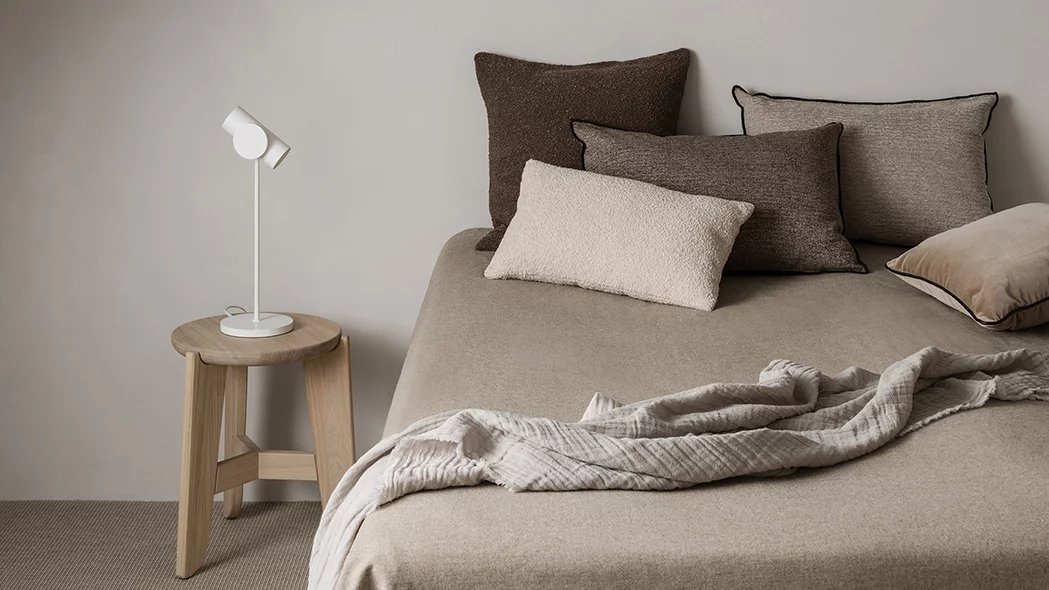 Eli Stool Sitzhocker von Blomus aus Holz neben einem Bett mit Kissen und Lampe.