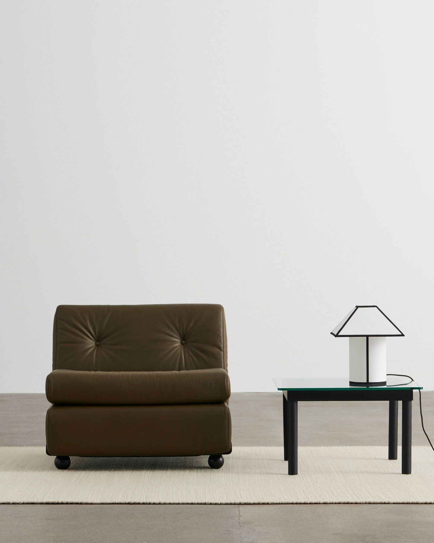 Ava Pyramid Table Lamp von Hay auf einem Tisch mit Glasplatte neben Sessel.