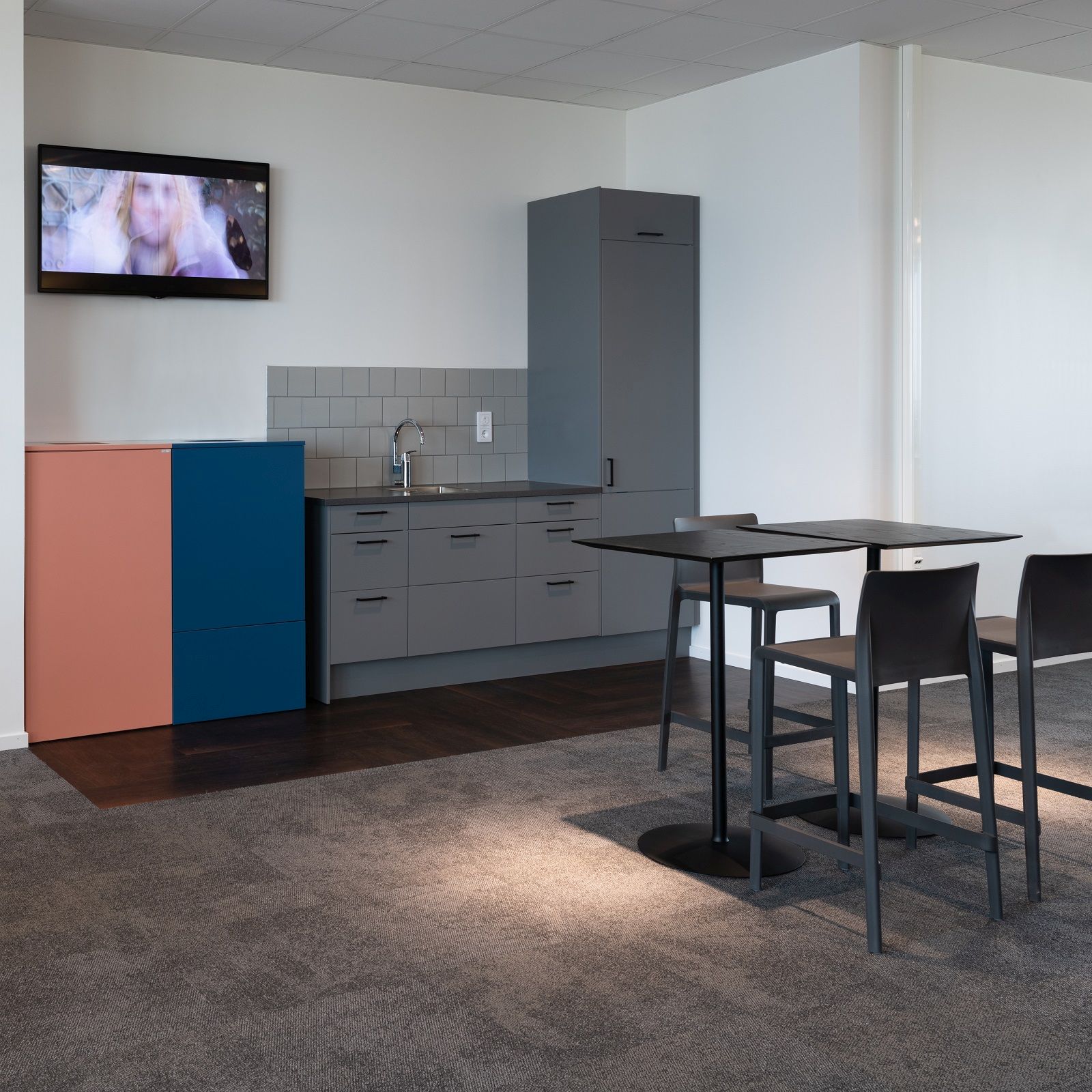 Büroküche mit farbigem Exhibit Bin Mülleimer zur Mülltrennung, grauen Küchenschränken und Stehtischen mit Barhockern.
