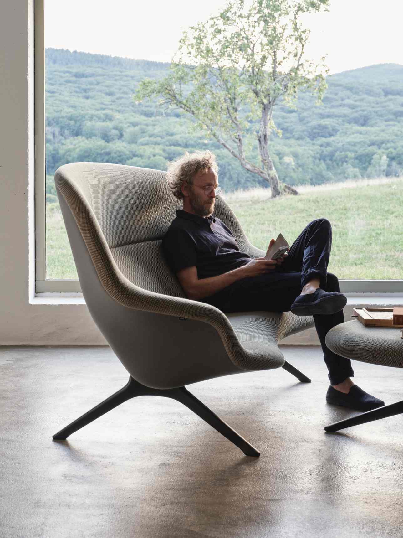 Mann liest in grauem Abalon Sessel von Vitra mit passendem Hocker vor Fenster.