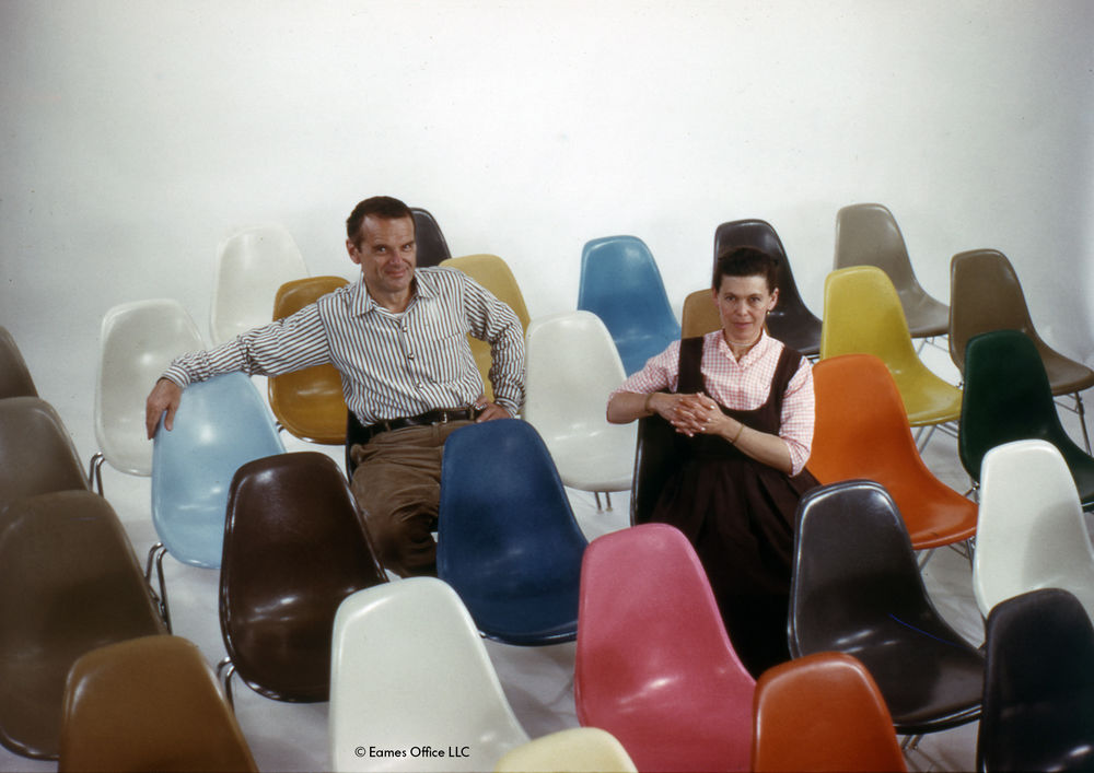 Charles und Ray Eames sitzen umgeben von bunten Eames Plastic Chairs, Designklassiker für Wohnzimmer und Büro.