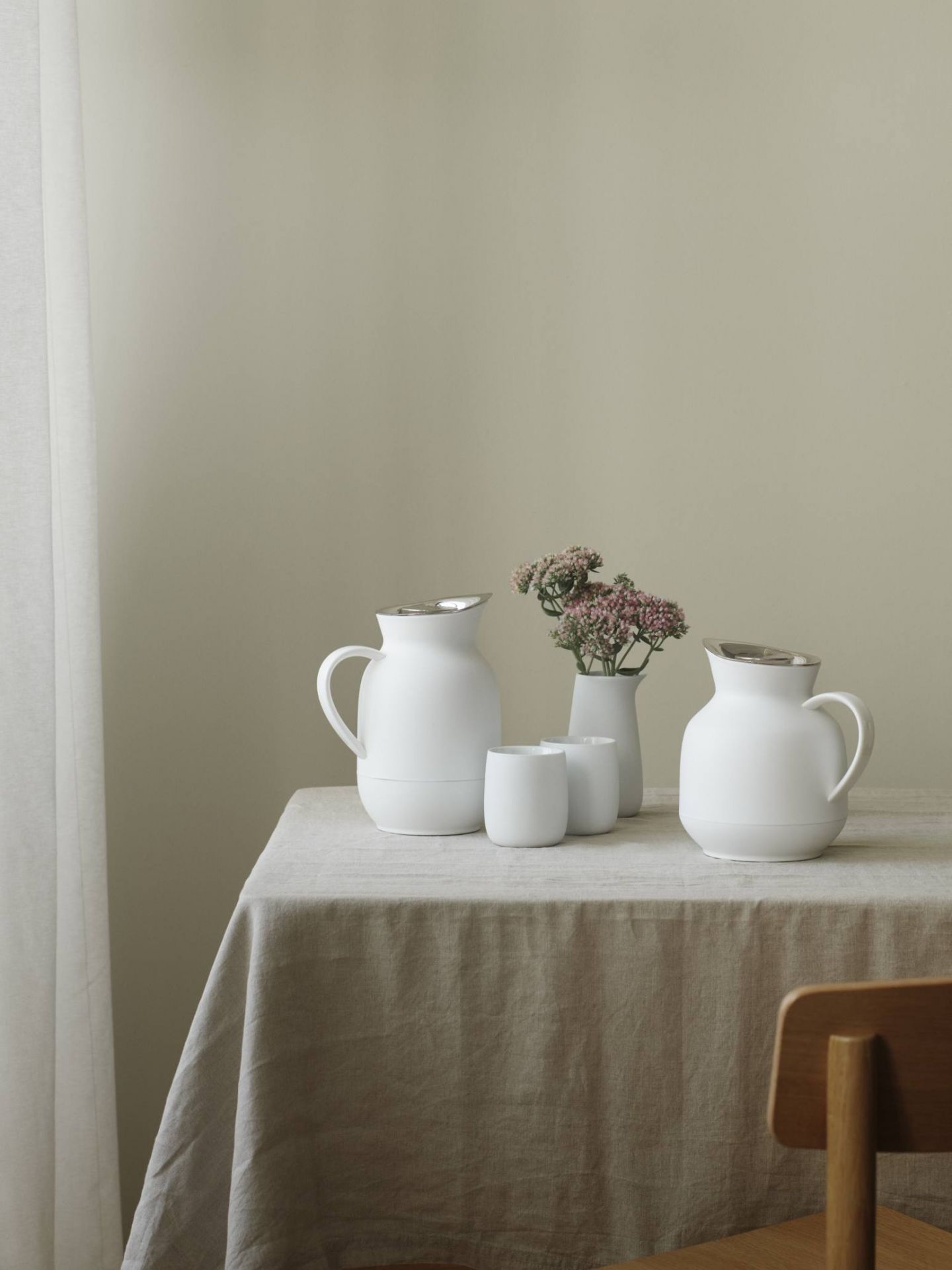 Stillleben mit weißen Keramik-Krügen, Bechern und Blumen auf einem Tisch mit Leinentuch.
