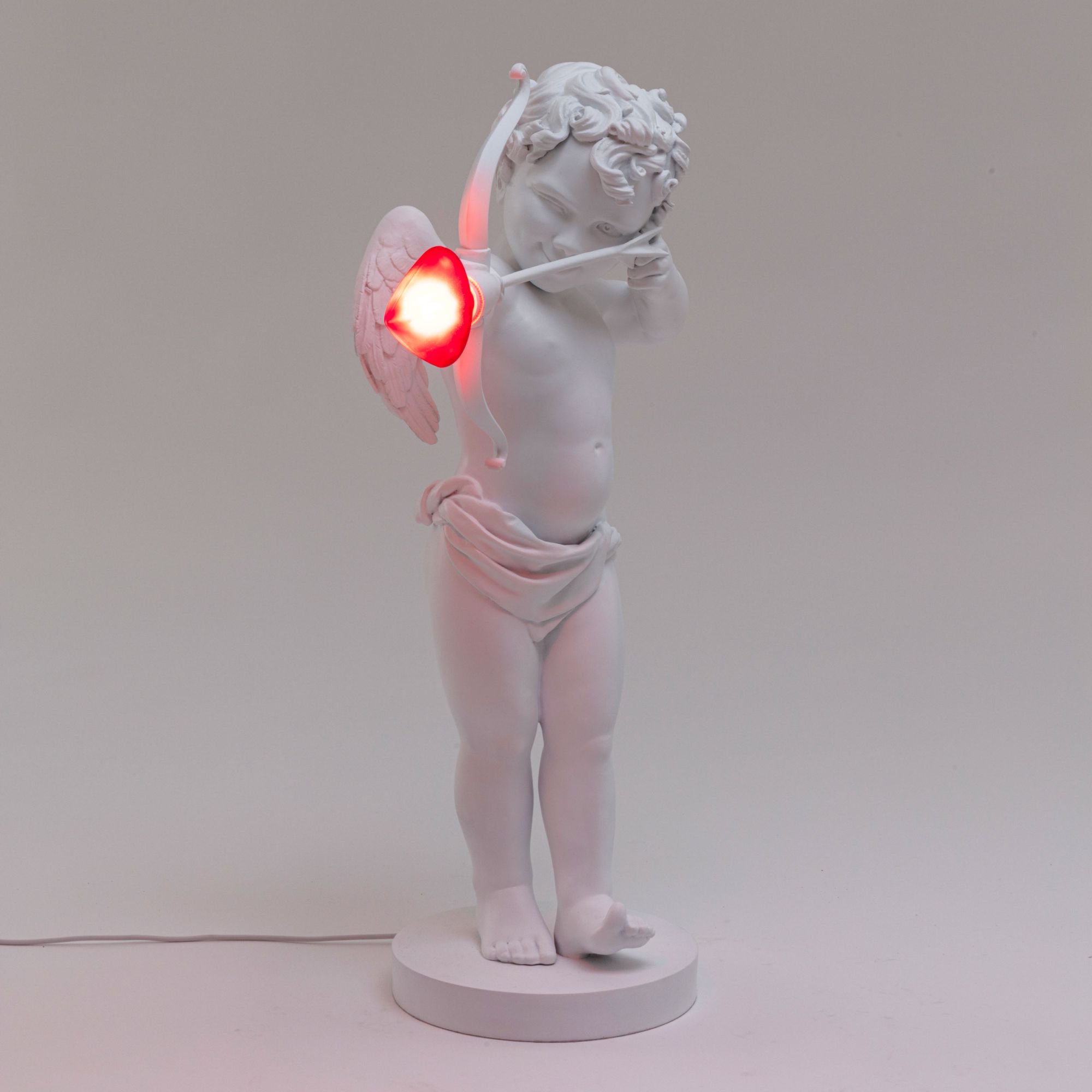 Cupid Lampe von Seletti: Tischleuchte in Form eines weißen Amor mit rotem Herz-Pfeil.
