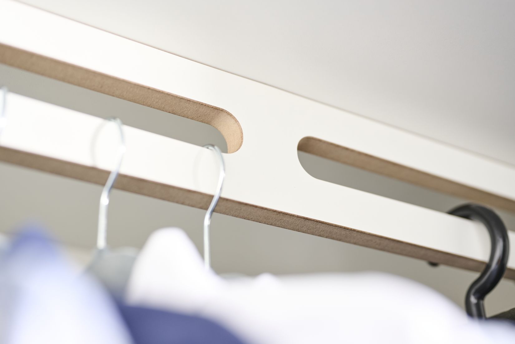 Detailaufnahme Tojo-Schrank Grundmodul: Weiße Schrankleiste mit Aussparungen für Kleiderbügel, Kleidung hängt darunter.
