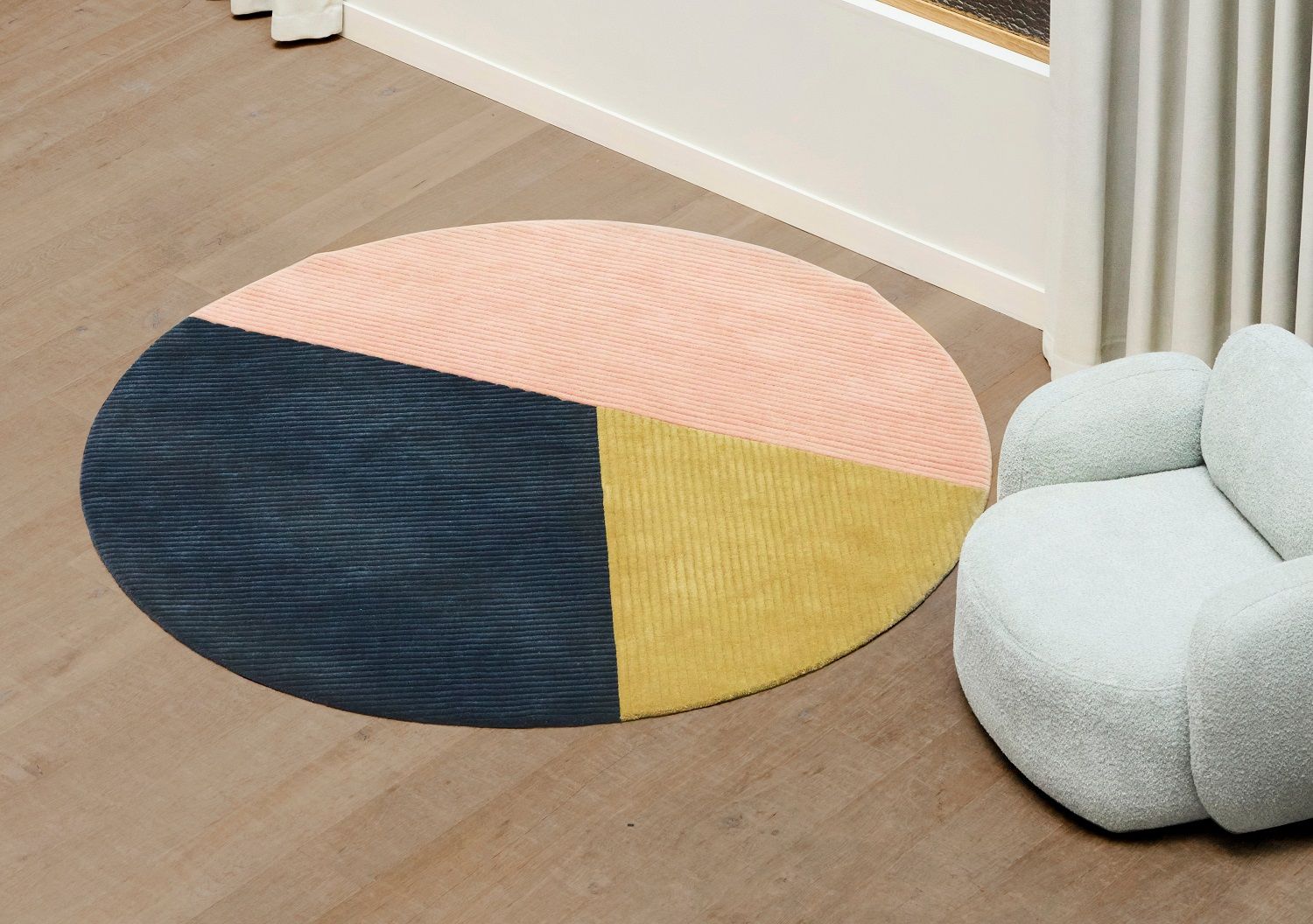 Runder Row Rug Teppich mit Farbblöcken in Rosa, Gelb und Dunkelblau auf Holzboden.