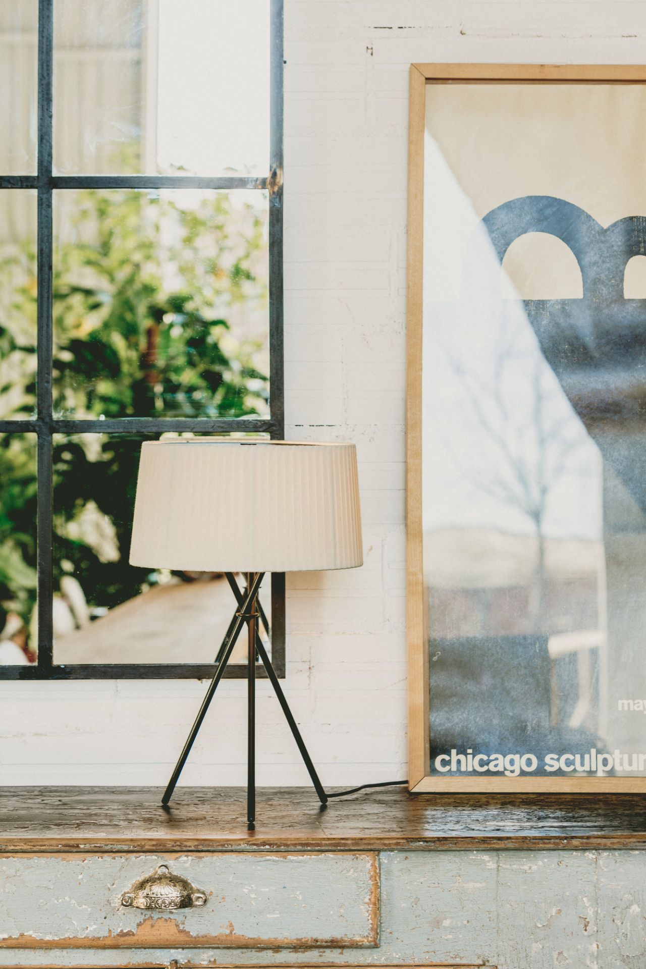 Beige Tischleuchte mit Stativfuß auf Vintage-Kommode, neben Fenster und gerahmtem Chicago Skulptur Poster.