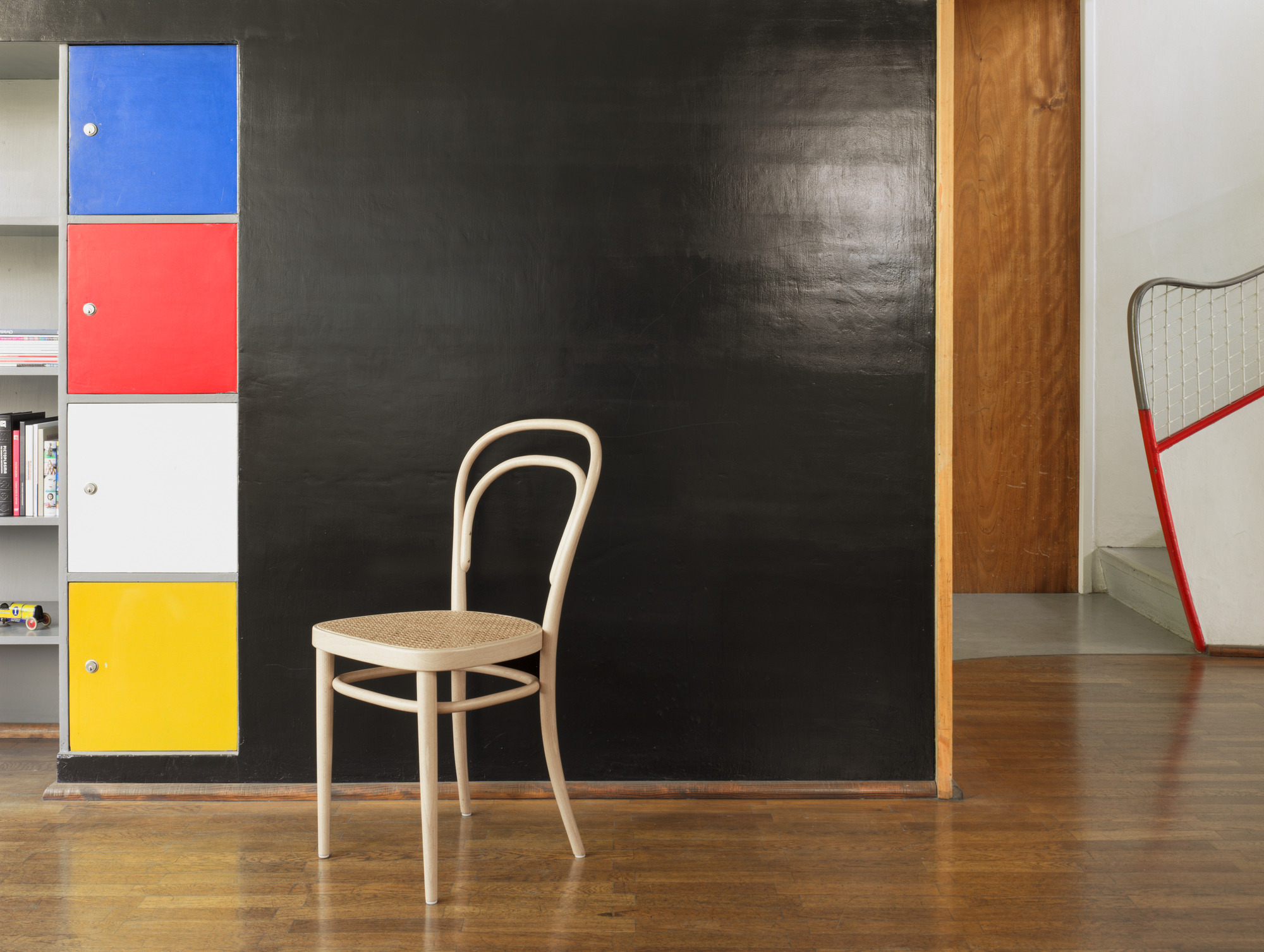 214 Bugholzstuhl, klassischer Wiener Kaffeehausstuhl mit Rohrgeflecht Sitzfläche vor schwarzer Wand und bunten Schränken.