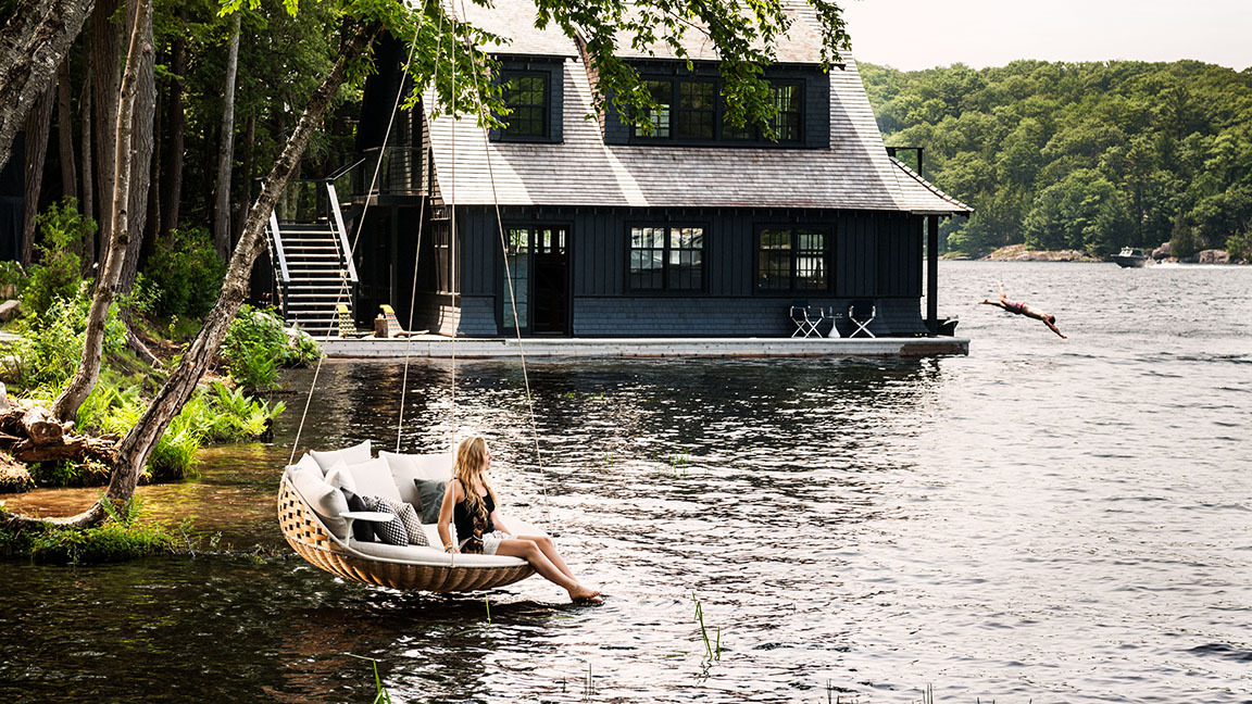 Frau entspannt auf der Dedon Swingrest Hängelounge mit Kissen am See, während eine Person ins Wasser springt.