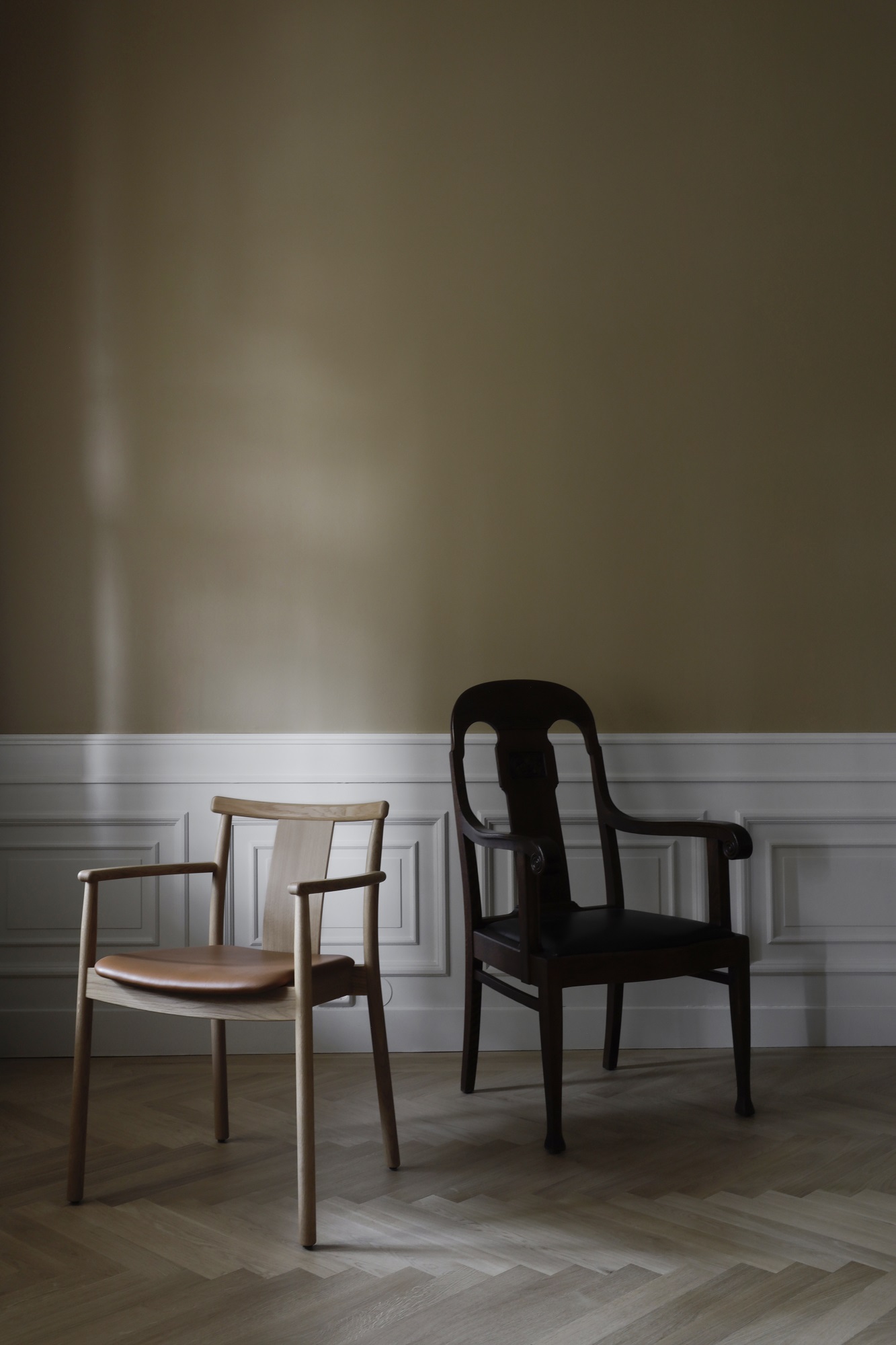 Zwei Esszimmerstühle: Heller Merkur Dining Chair mit Armlehnen und dunkler Holzstuhl vor Wandpaneelen.
