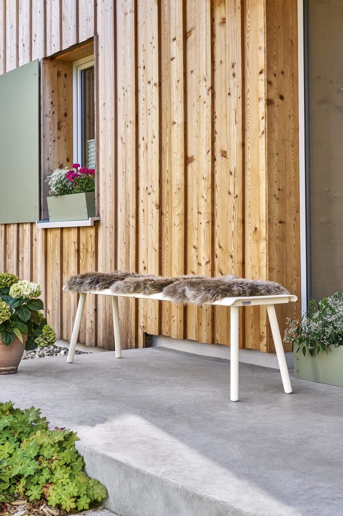 Weishäupl Vapio Bank für den Außenbereich mit Fellauflage vor einer Holzwand und Blumenkästen.