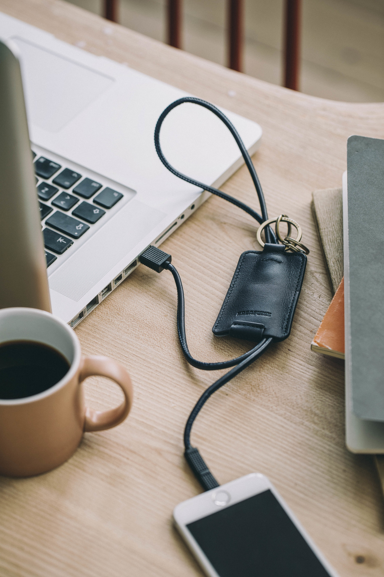 cCHAIN Schlüsselanhänger mit USB-Kabel von Kreafunk lädt Smartphone am Laptop, Kaffeetasse daneben.