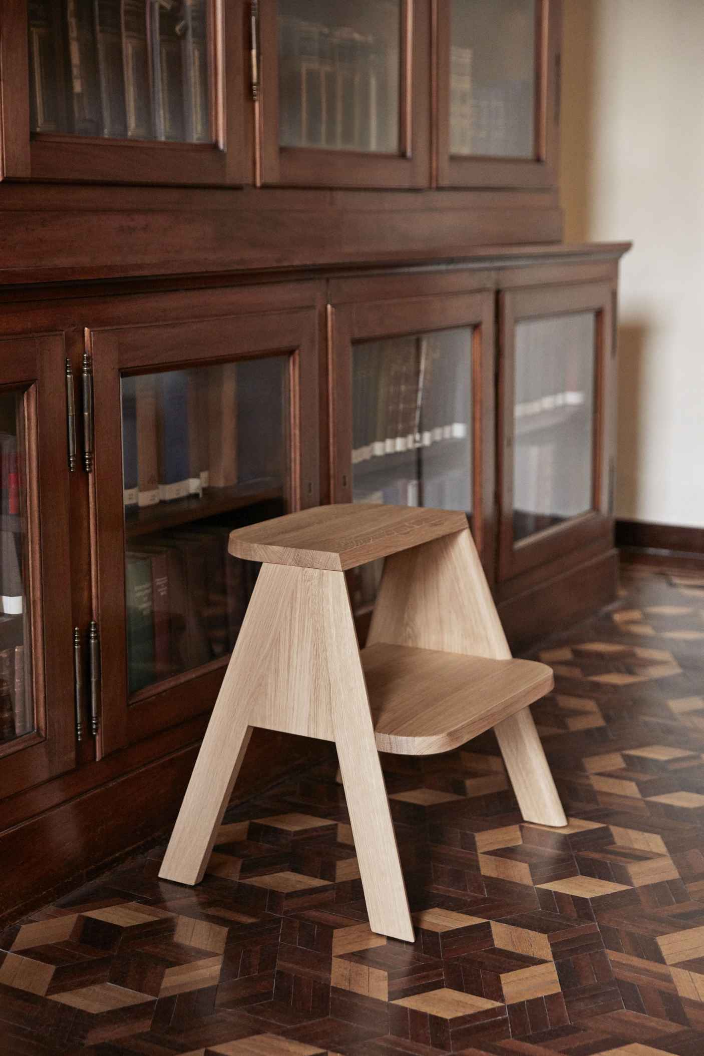Butler Hocker von Hay aus hellem Eichenholz vor einem dunklen Holzschrank und Parkettboden.