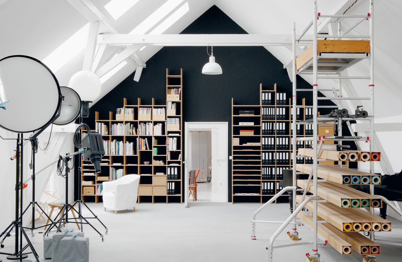 Helles Dachgeschoss-Büro mit Regalen, Büchern, Ordnern, Fotostudio-Equipment und einer fahrbaren Arbeitsbühne.