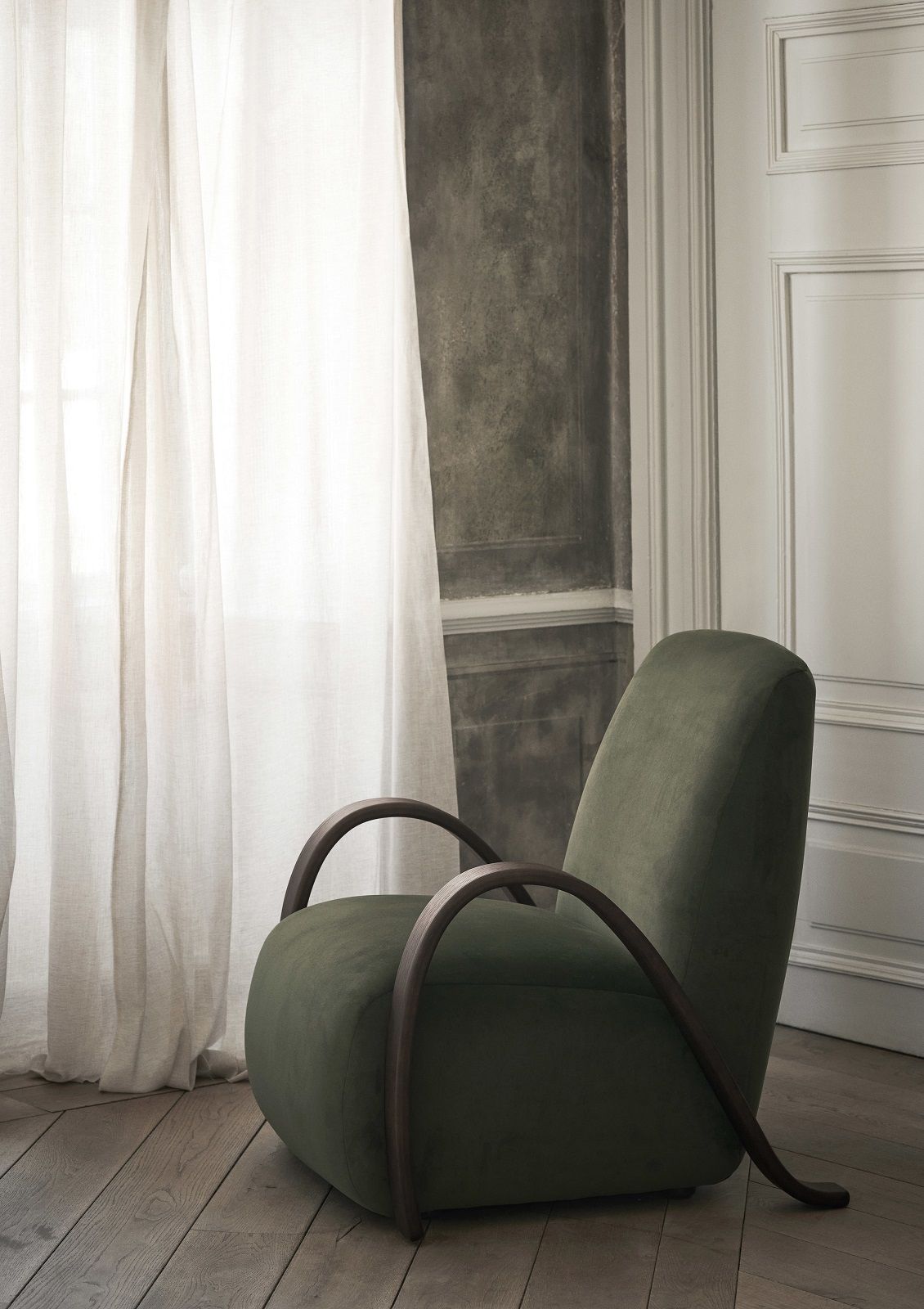 Grüner Buur Lounge Chair Sessel von Ferm Living mit Holzrahmen vor Vorhang und Wand.
