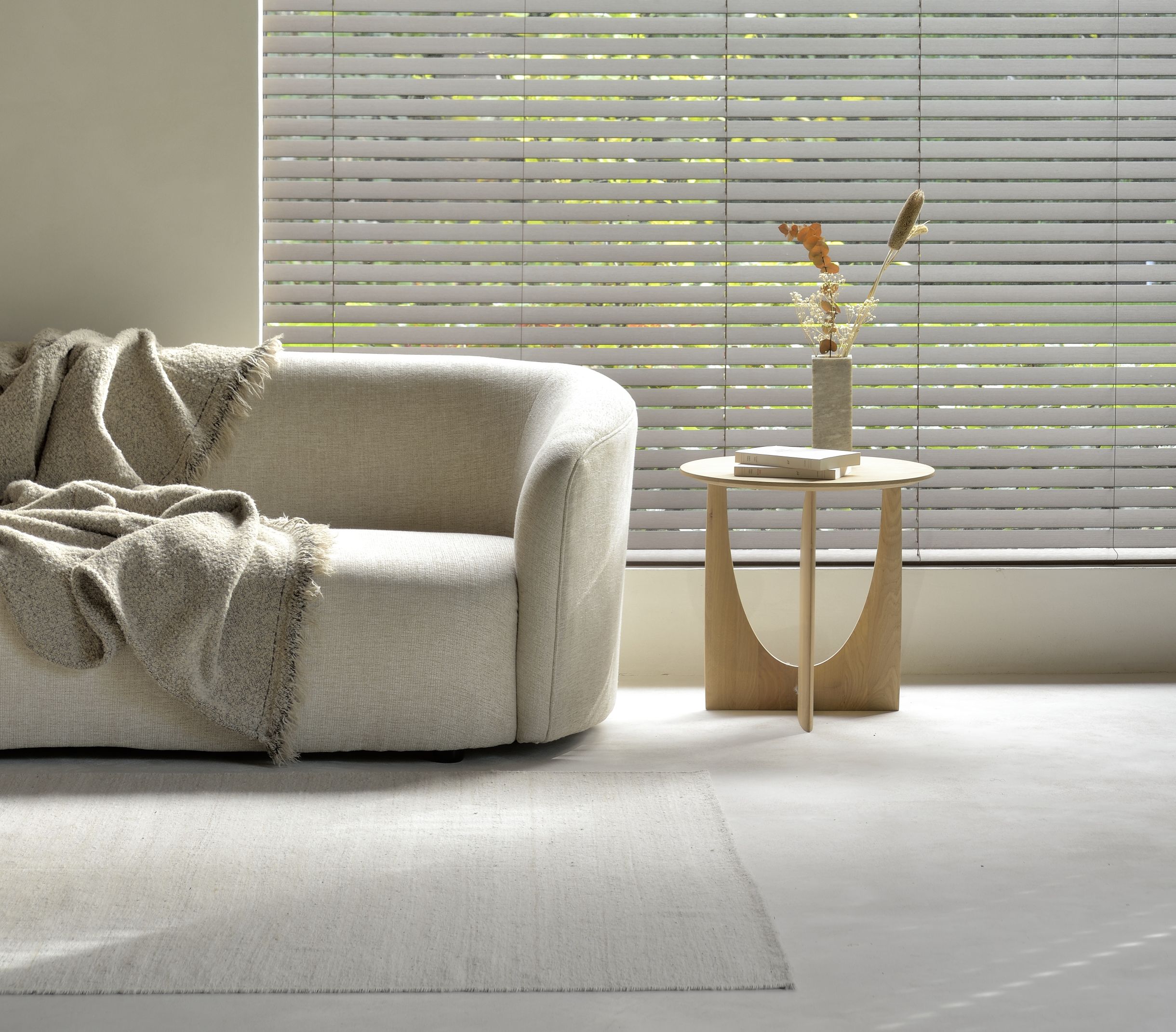 Beige Couch mit Decke, Holztisch mit Dekoration und heller Teppich vor Fensterjalousien.