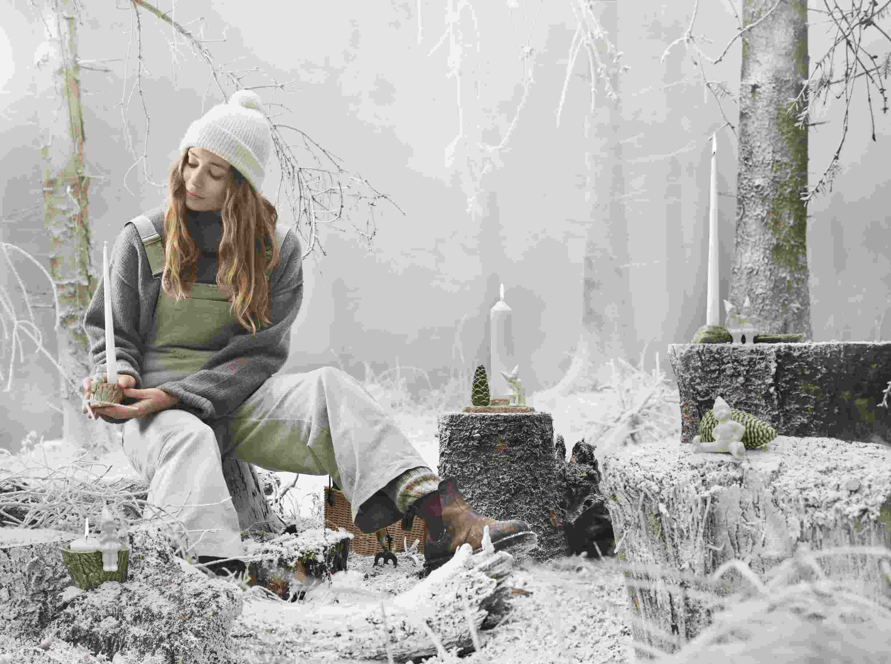 Frau im Winterwald mit Kähler Christmas Tales Julekram Kerzenständer und weiterer Weihnachtsdekoration auf Baumstümpfen.