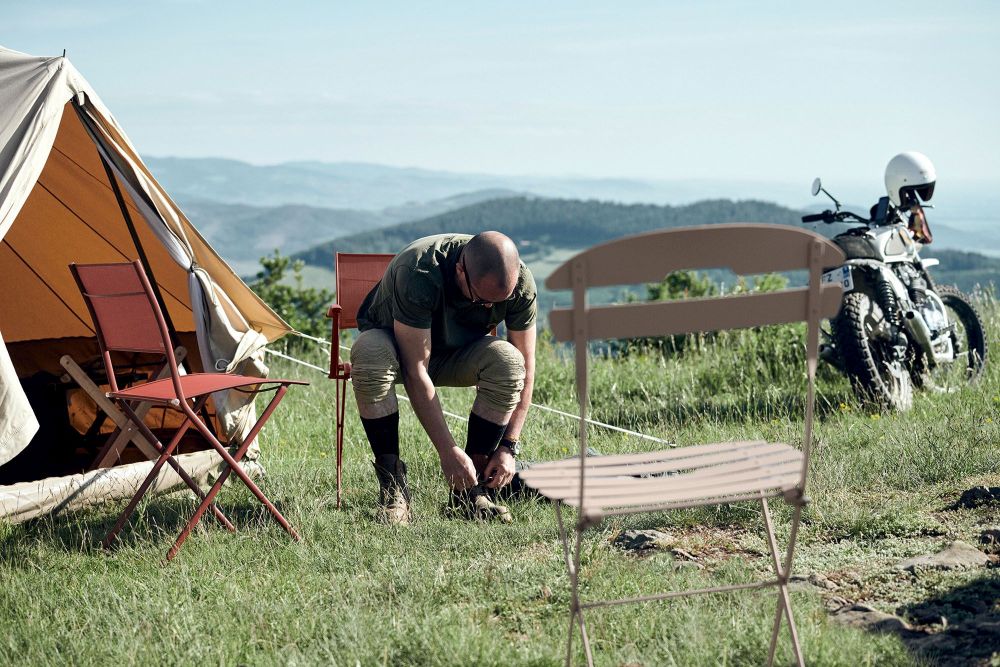 Camping-Szene mit La Môme Klappstühlen von Fermob, Mann bindet Schuhe, Zelt, Motorrad und Berglandschaft.