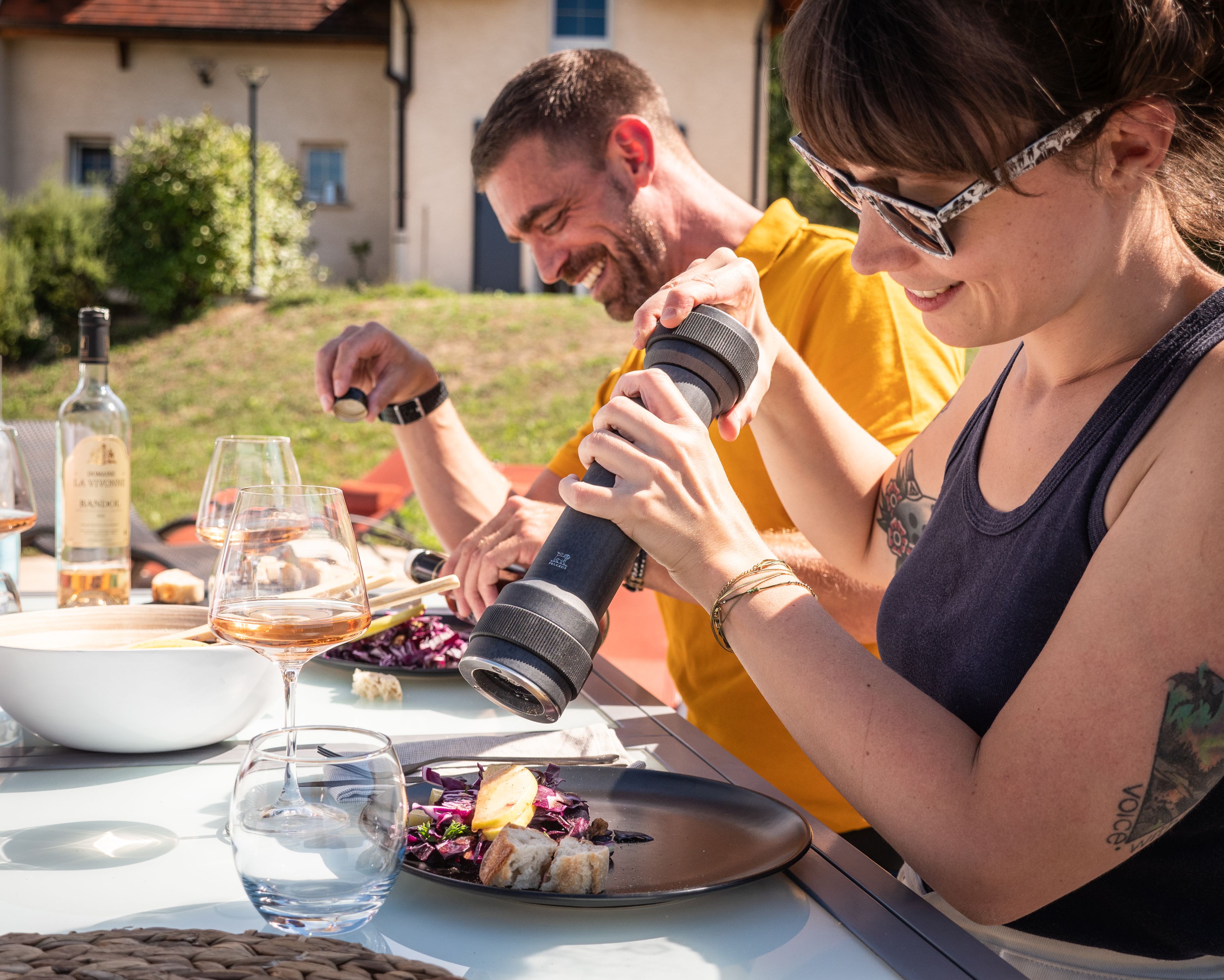 Draufsicht: Frau würzt Salat mit Peugeot BBQ Pfeffermühle graphit, Outdoor-Szene mit Wein.