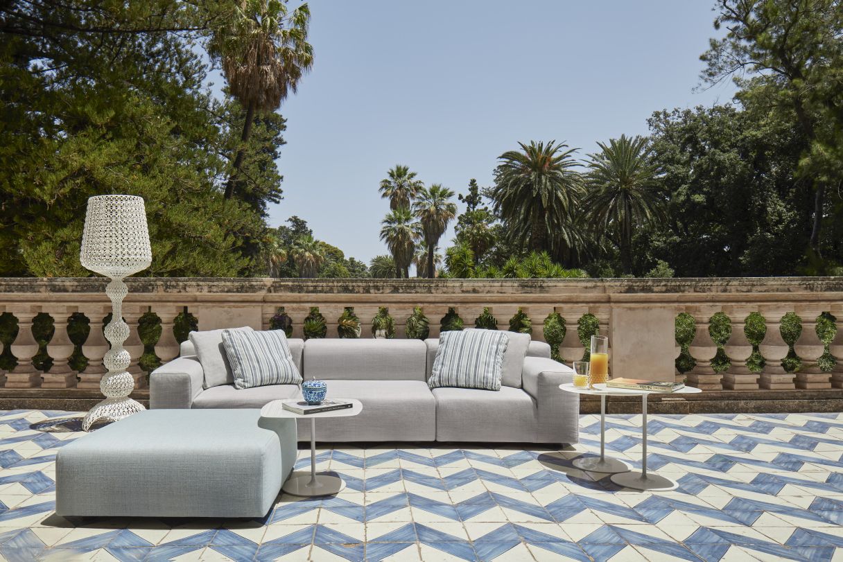 Graues Kartell Outdoor Sofa mit Kissen und Beistelltischen auf einer Terrasse mit blau-weißem Boden.