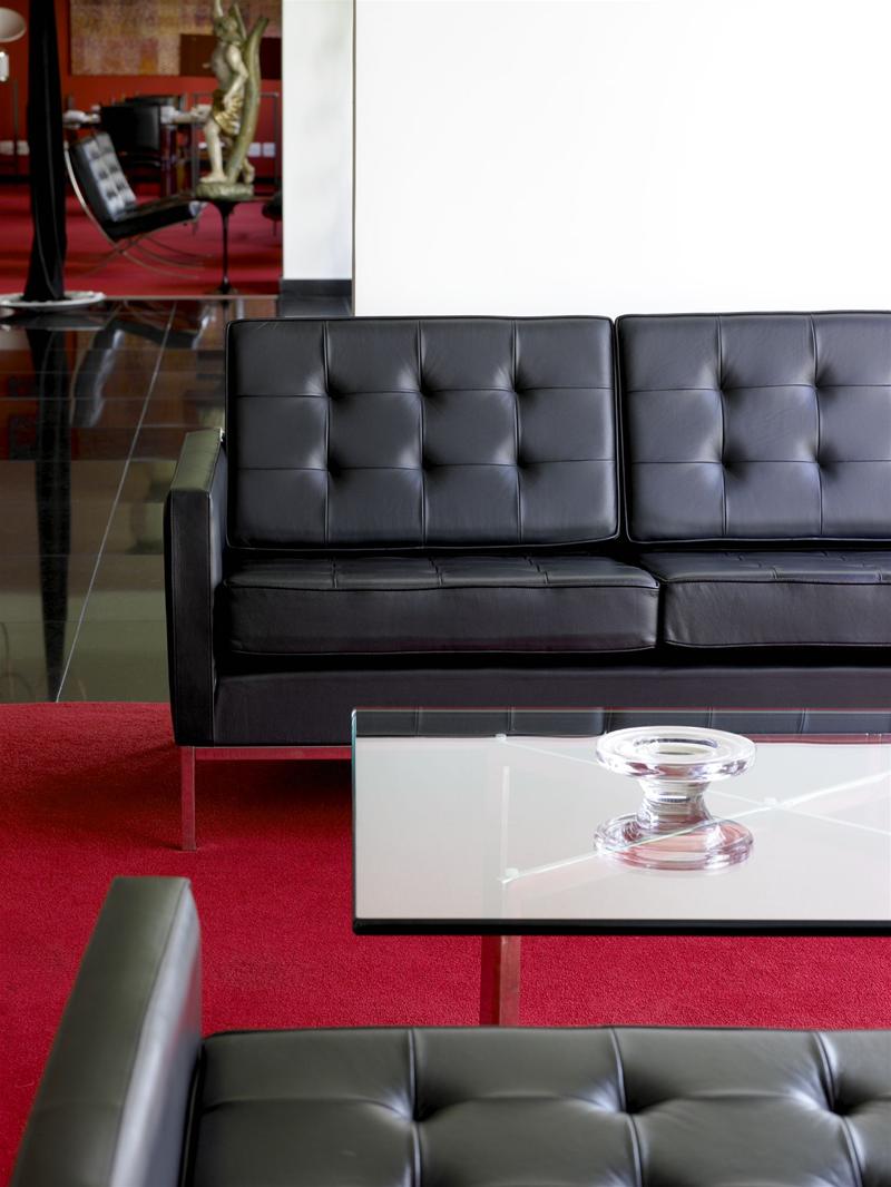Barcelona Couchtisch aus Kristallglas von Knoll International neben schwarzem Ledersofa auf rotem Teppich.