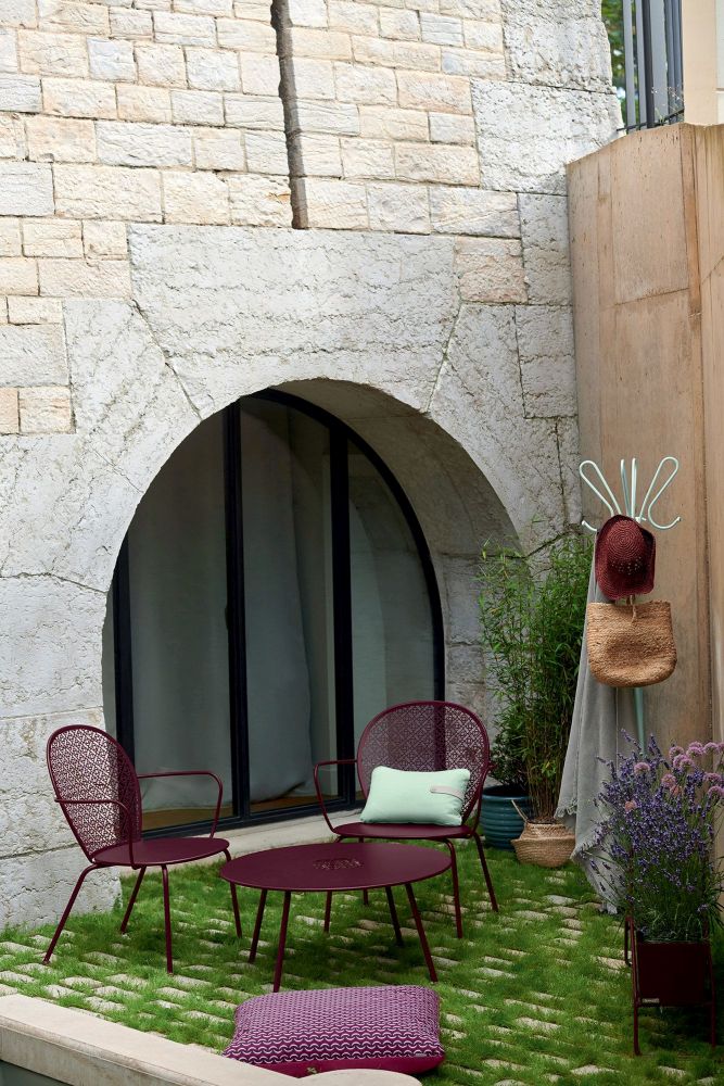 Fermob Lorette Sessel und Tisch in Pflaume auf Rasenfläche vor Steinmauer. Outdoor-Möbel für Garten und Terrasse.