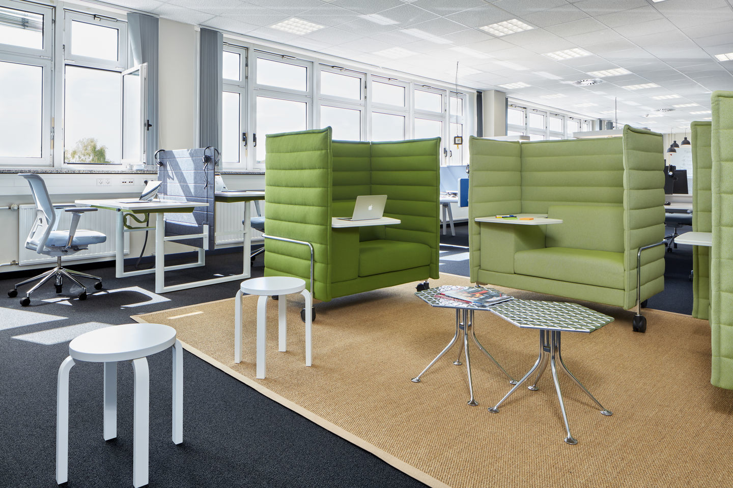 Büro mit Vitra ID Mesh Bürodrehstuhl, grünen gepolsterten Arbeitsinseln und weißen Hockern für moderne Büroeinrichtung.