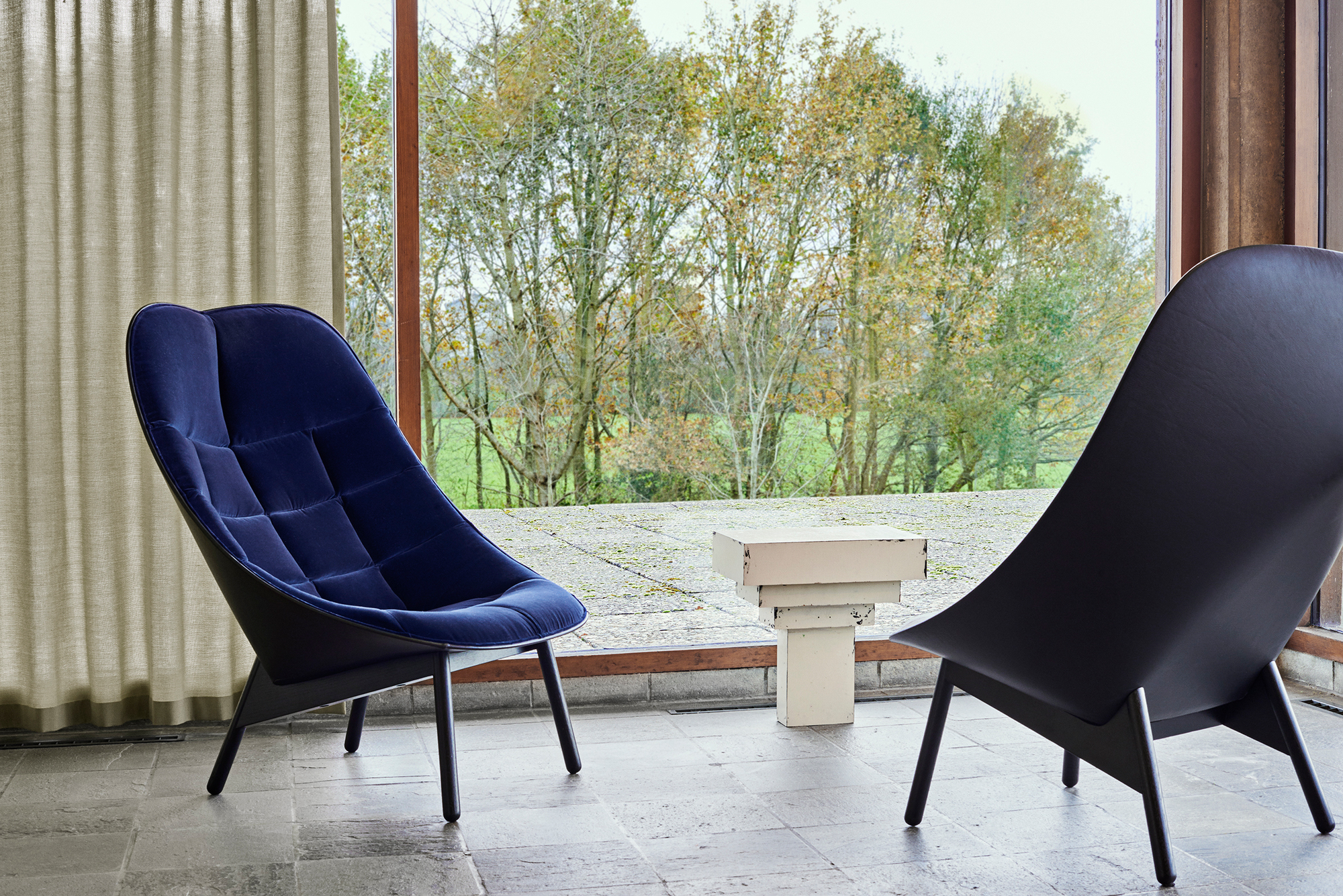 Blauer Uchiwa Quilt Sessel von Hay und ein schwarzer Sessel vor einem Fenster mit Baumblick.