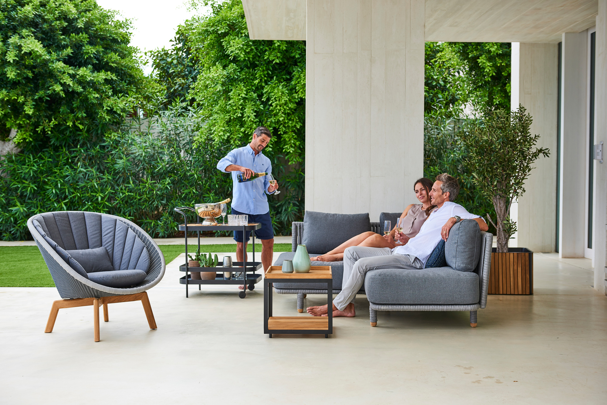 Peacock Loungesessel in Schwarz von Cane-Line mit Aluminiumbeinen im Garten mit entspannten Personen.