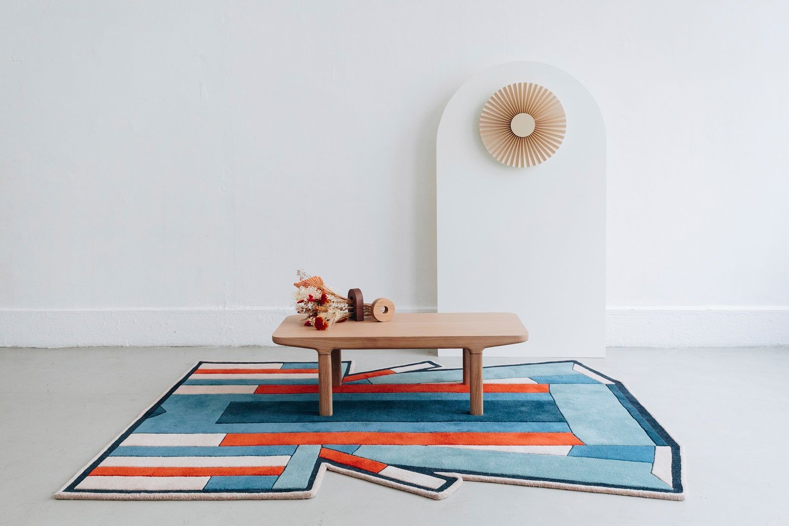 Inès Carpet Teppich Hart™: Moderner Teppich mit geometrischen Mustern in Blau, Orange und Weiß unter einem Holztisch.
