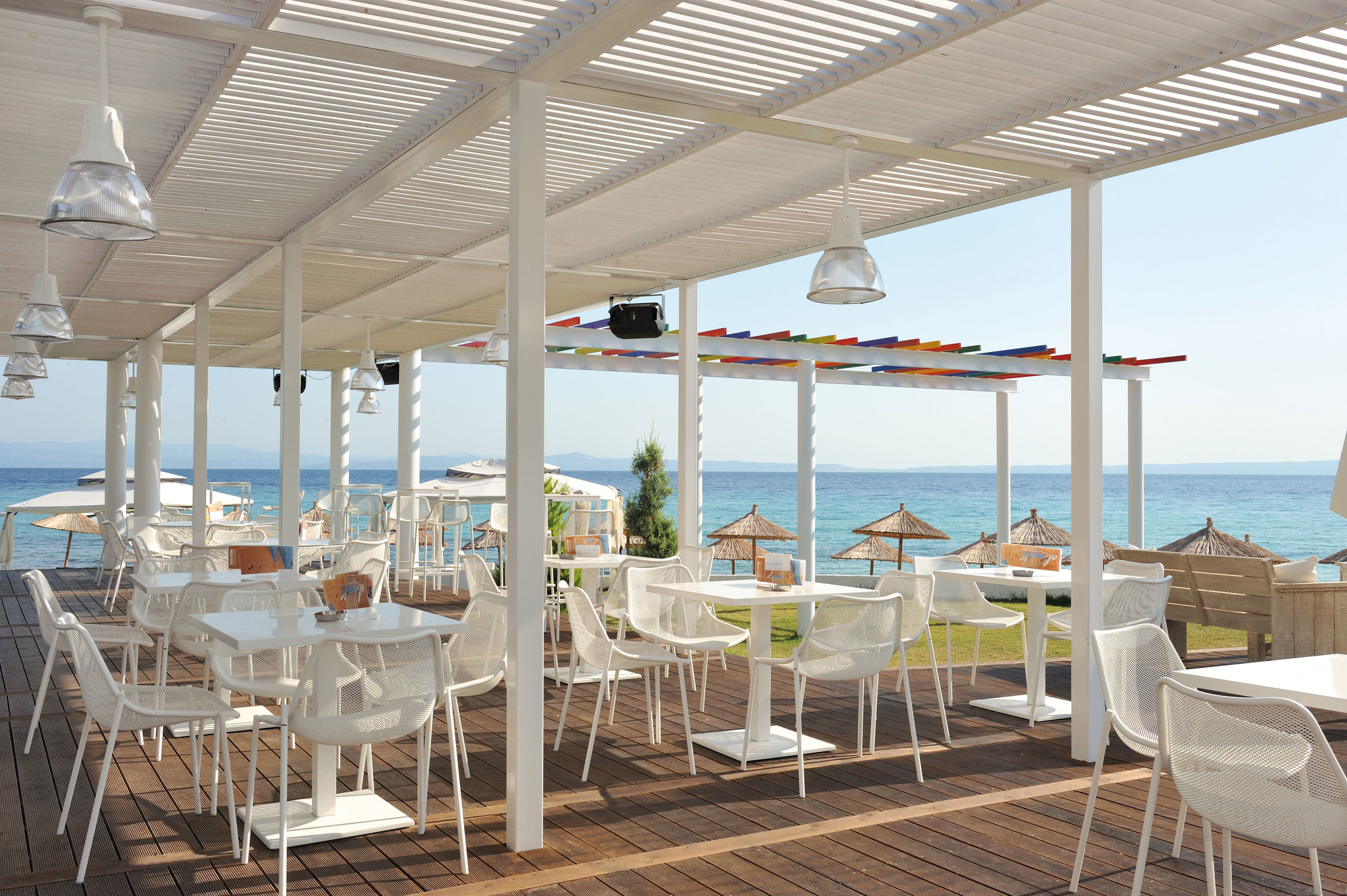 Außenbereich mit weißen Emu Stühlen und Tischen auf einer Holzterrasse am Meer.