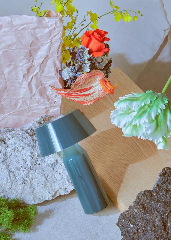 Blaue Bicoca Akkuleuchte von Marset als Tischleuchte, umgeben von Blumen und Steinen.