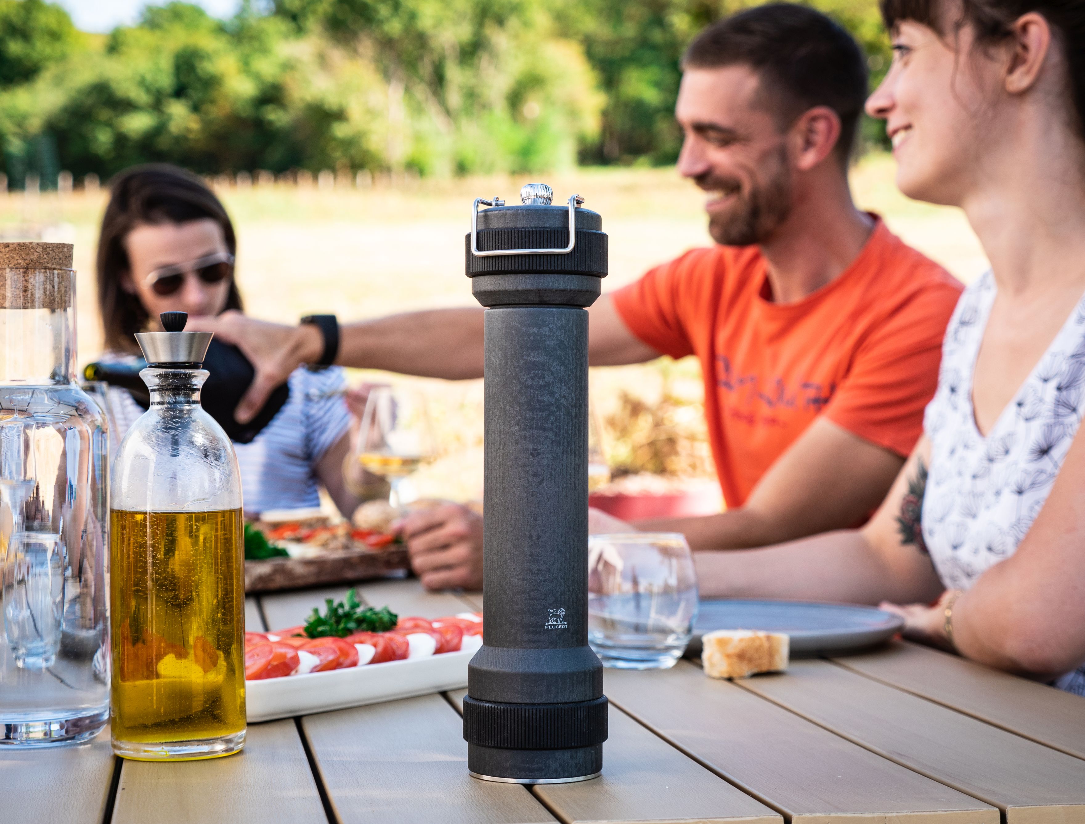 Peugeot BBQ Pfeffermühle Graphit 30 cm auf Tisch mit Familie beim Grillen im Freien.