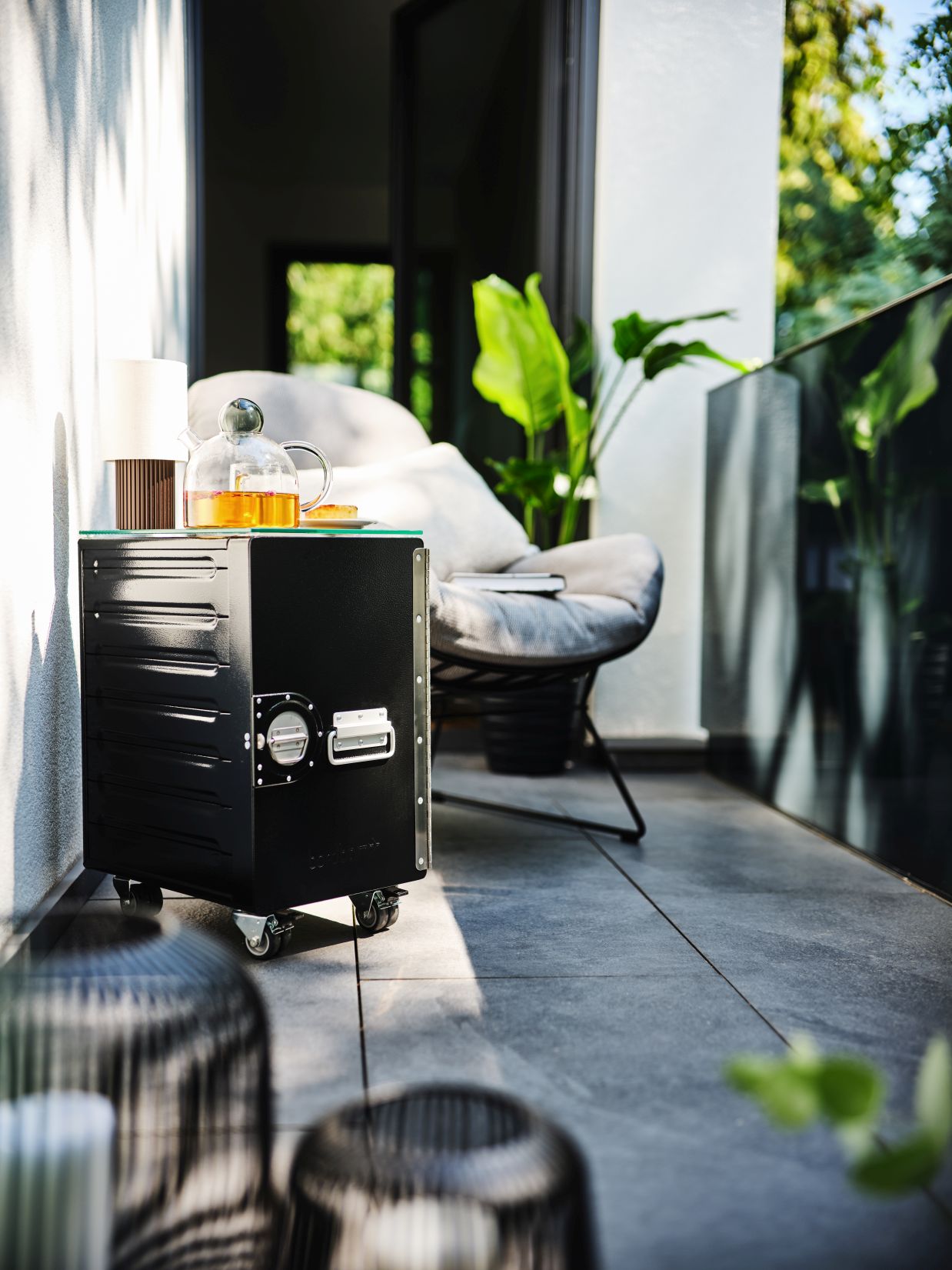 Schwarzer Box Rollwagen Bordbar mit Glasplatte auf Balkon, Teekanne und Lampe darauf. Mobiler Servierwagen.