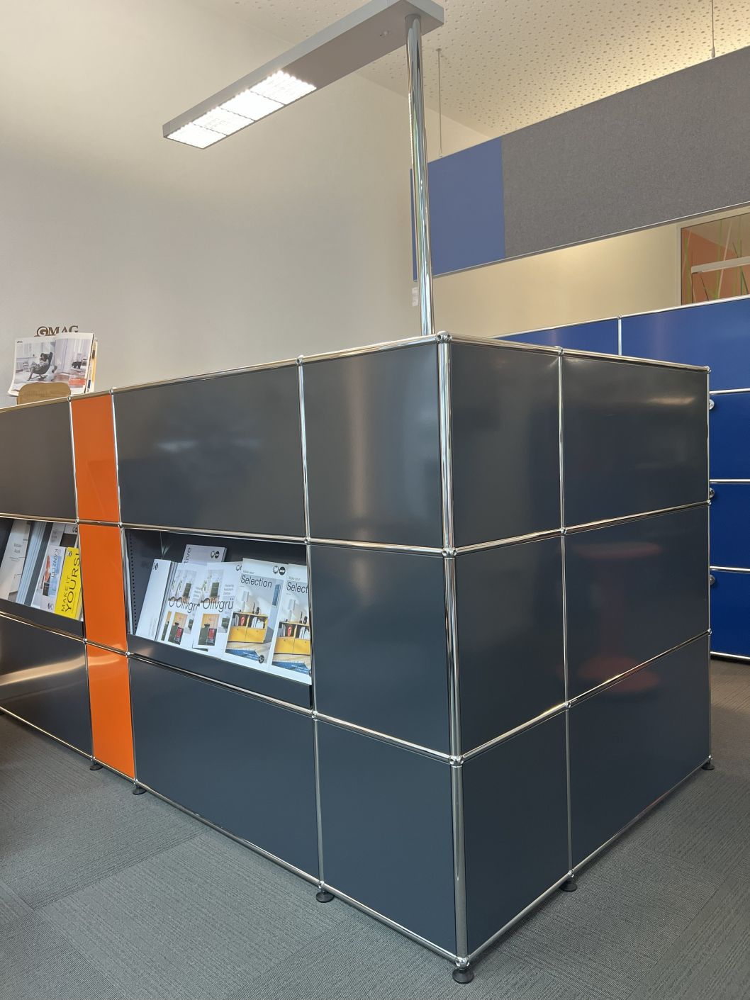 USM Haller Theke, Ausstellungsstück in Anthrazit und Orange, mit integrierten Regalen für Zeitschriften und Katalogen.