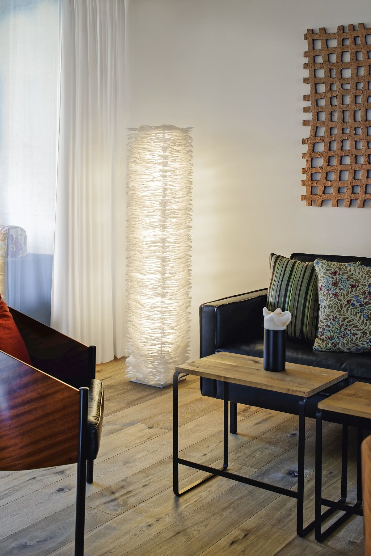 Moderne Wohnzimmergestaltung mit weißer Design-Stehlampe, schwarzem Ledersofa und Holztischen auf hellem Parkettboden.