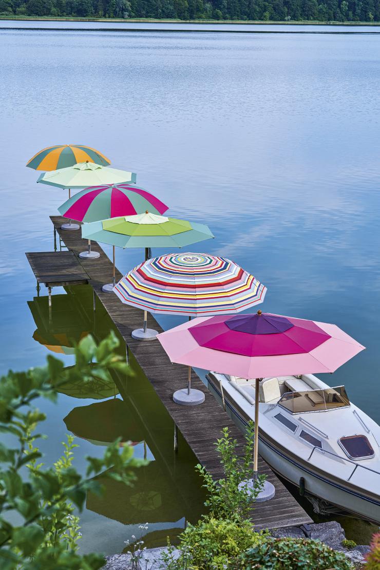 Mehrere bunte Weishäupl Sonnenschirme/Pagodenschirme auf einem Holzsteg am See, neben einem Boot.