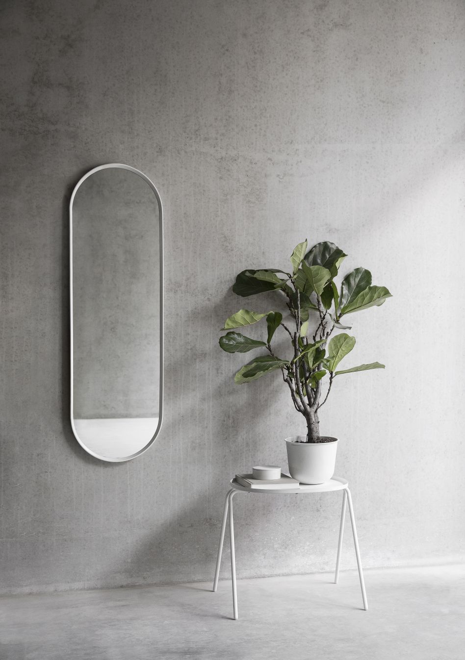 Norm Wandspiegel Oval von Audo mit weißem Rahmen an grauer Wand, neben Pflanze.