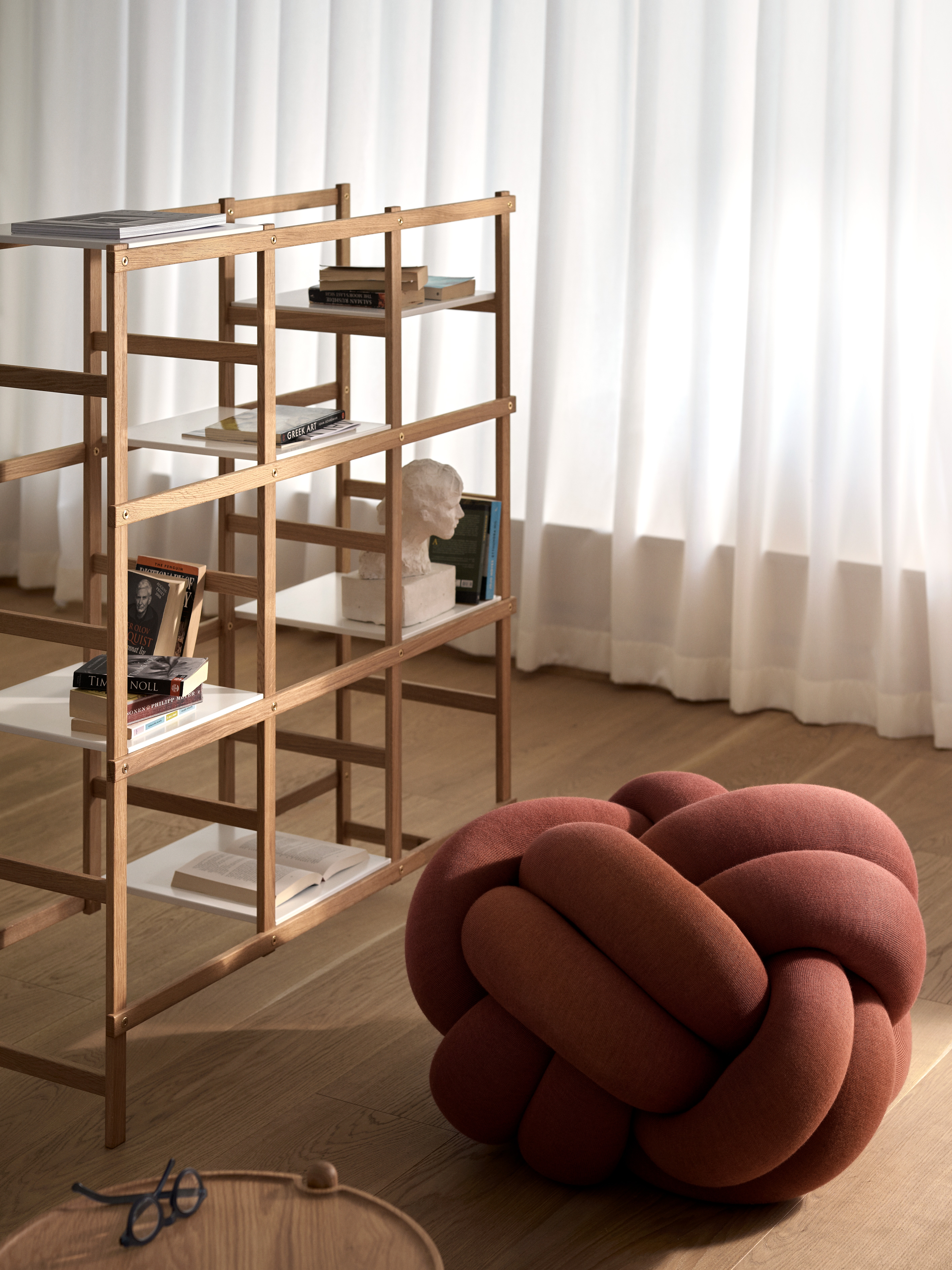 Helles Wohnzimmer mit Holzregal, Bücher, Dekoration und rotem Knotenkissen auf Parkettboden.