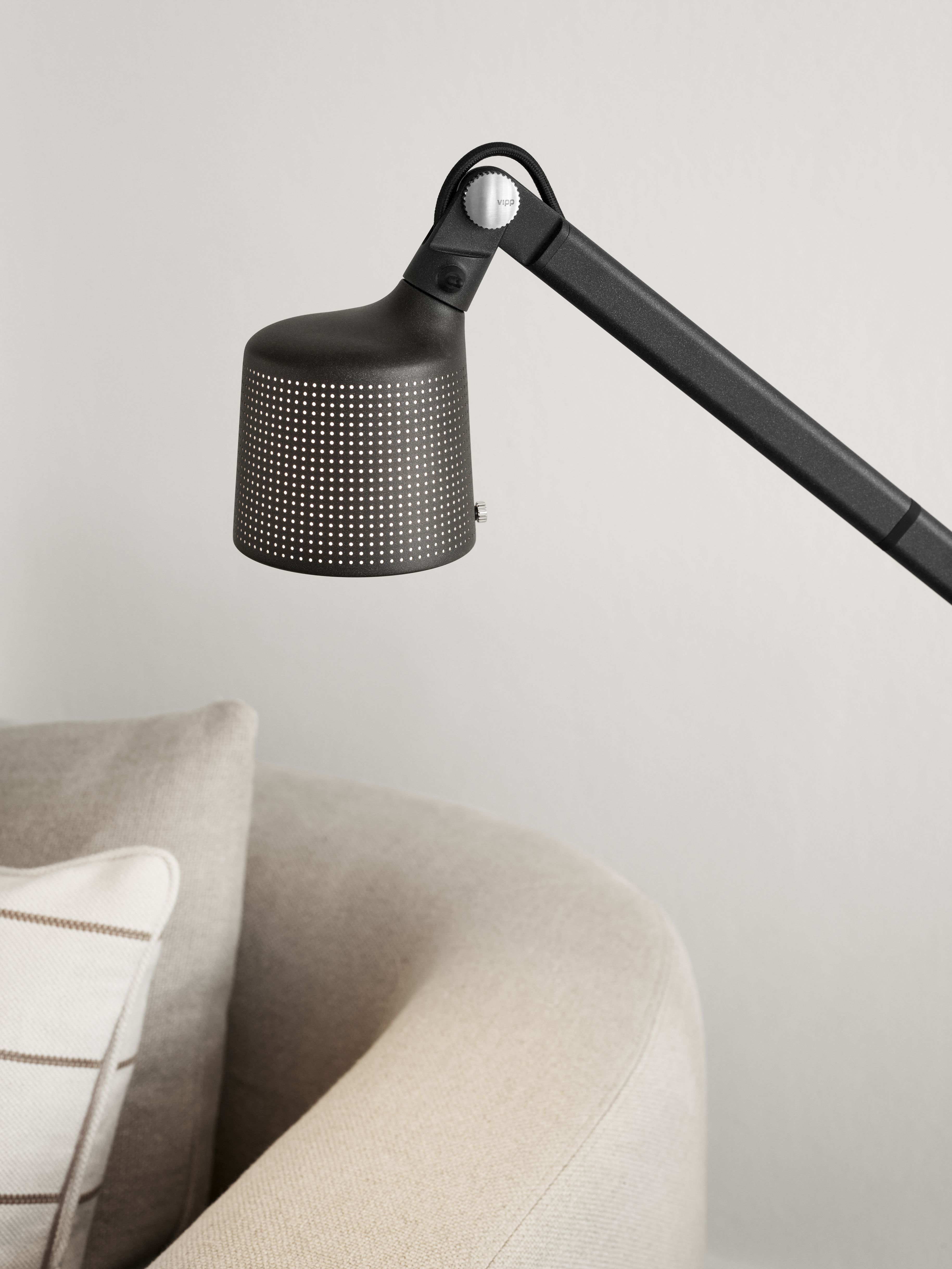 Schwarze Vipp Boden Leselampe mit verstellbarem Arm und perforiertem Lampenschirm neben einem beigen Sessel.