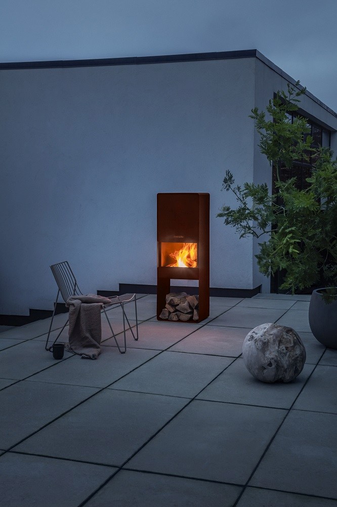 Eva Solo FireBox Garden Holzkamin mit brennendem Feuer und Holzscheiten auf einer Terrasse.