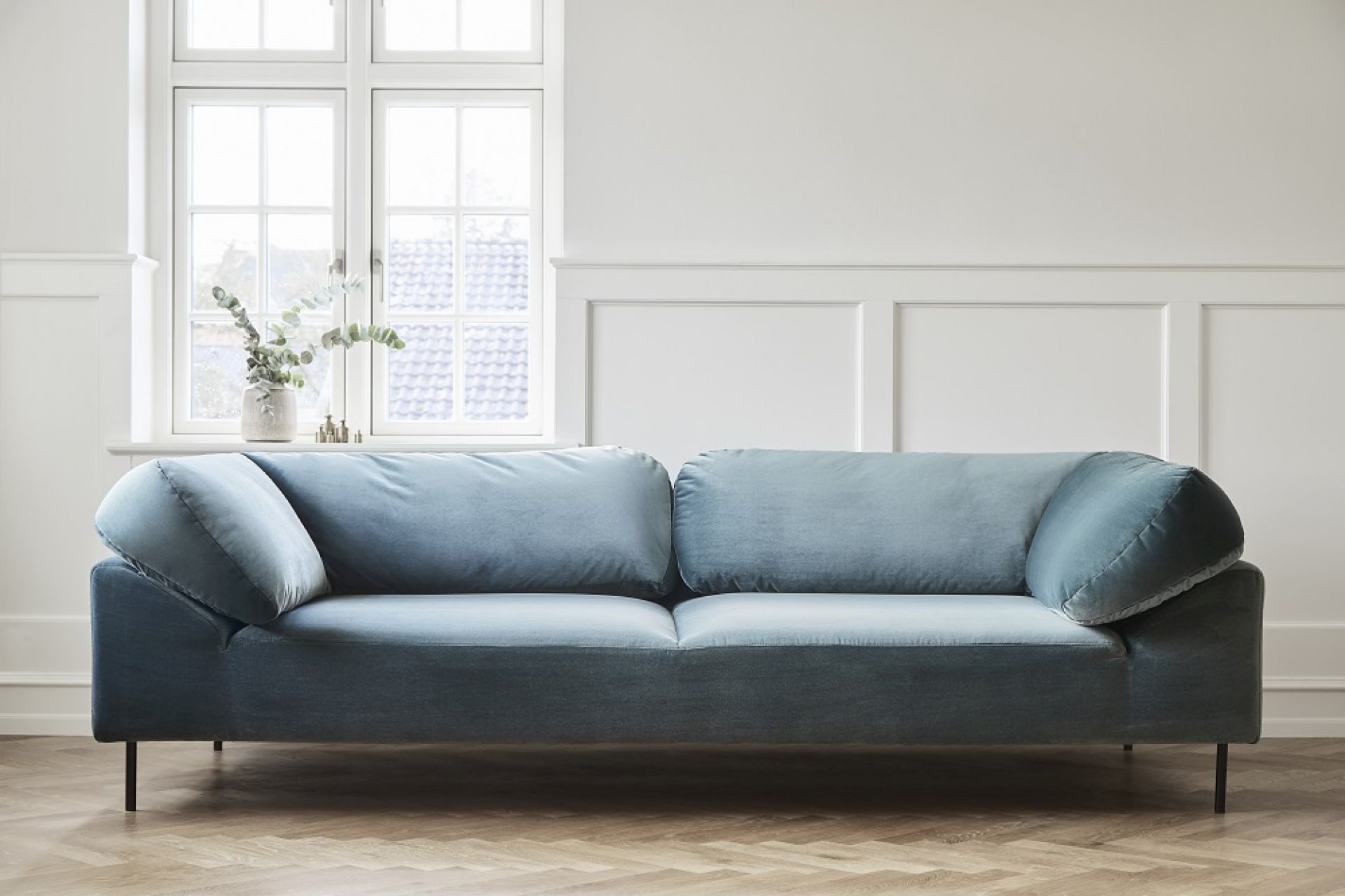 Hellblaues, modernes Sofa mit Kissen und schwarzen Füßen vor weißer Wand und Fenster.