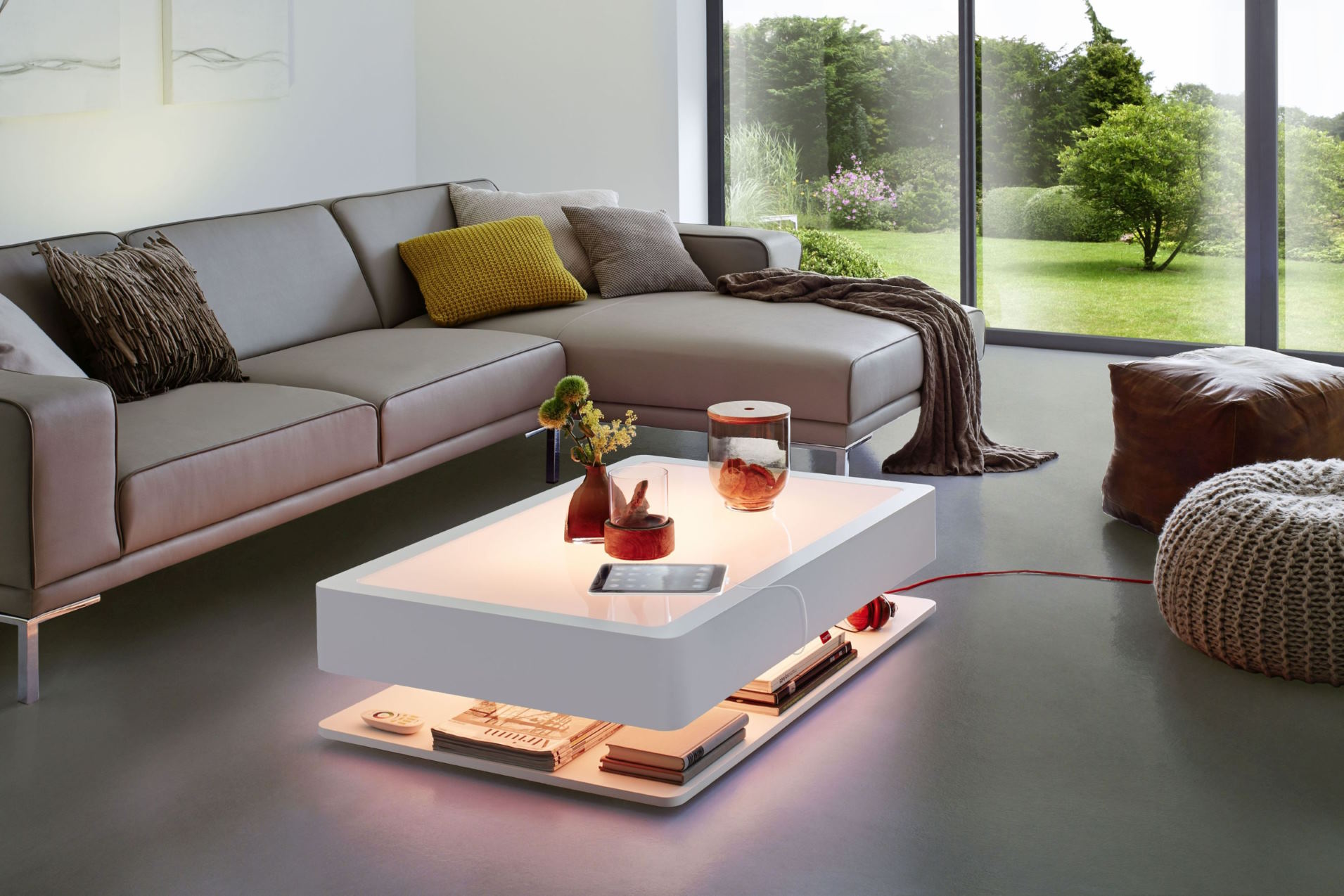 Moderner Wohnraum mit beleuchtetem Couchtisch, grauer Ledercouch, Kissen und Blick in den Garten.