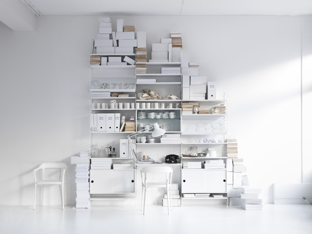 Helles Wohnzimmer mit weißem String Furniture Regalsystem, Schreibtisch und Stühlen, gefüllt mit Dekoration und Büromaterial.