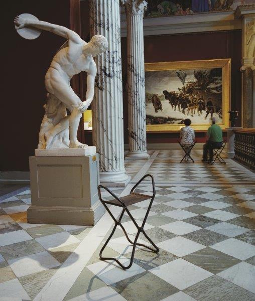 Schwarzer Klapphocker aus Metall und Rattan im Museum, neben Statue und Marmorsäulen auf Schachbrettmuster-Boden.