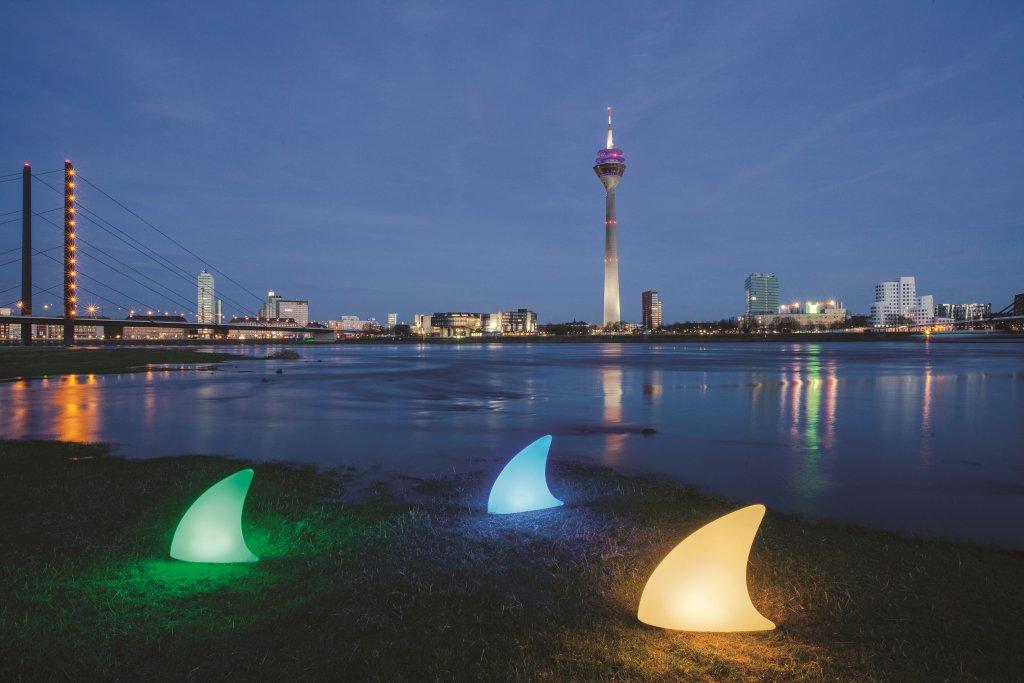 Drei leuchtende Moree Haifischflossen Bodenleuchten in Grün, Blau und Gelb am Ufer mit Skyline im Hintergrund.