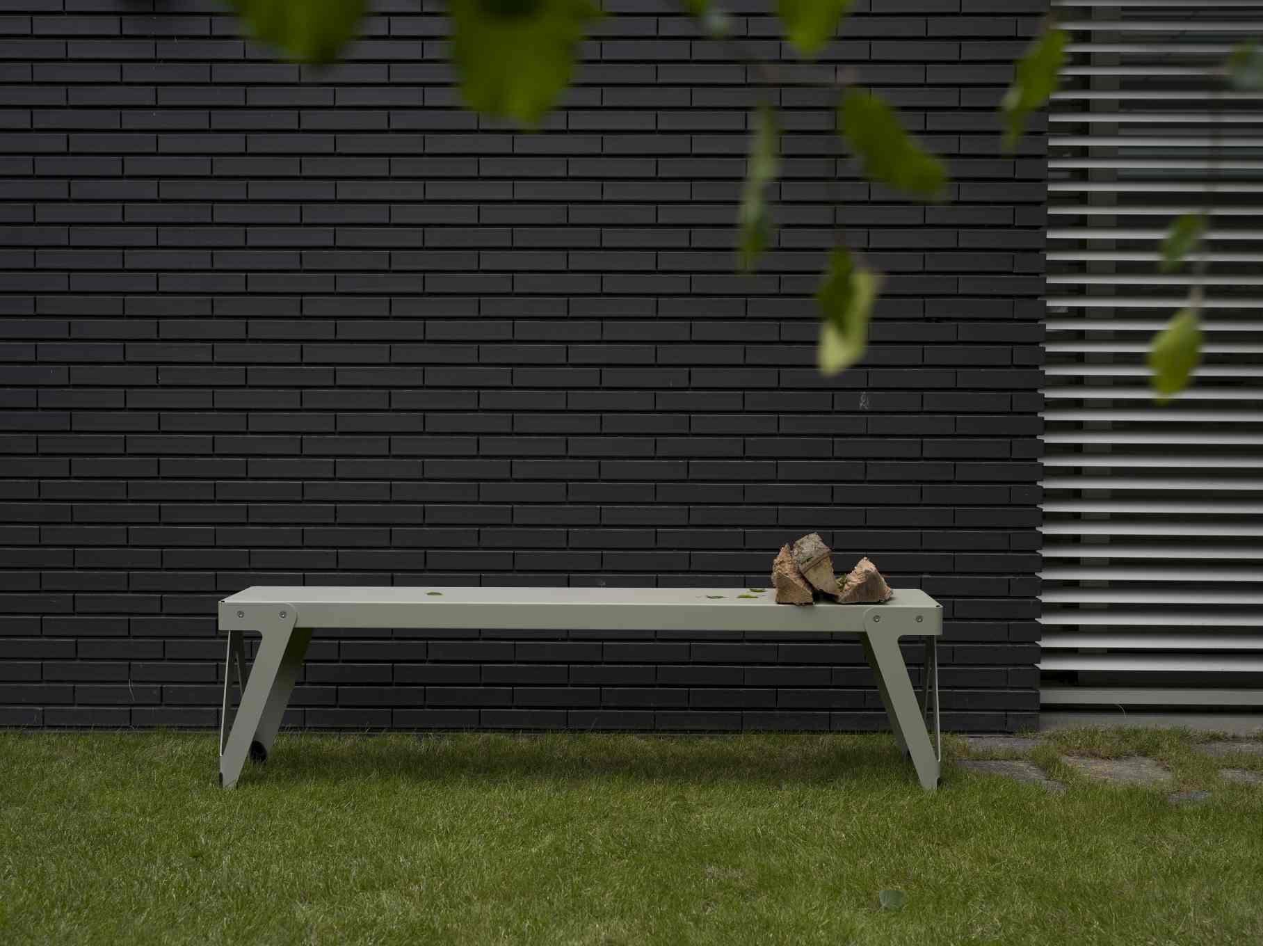 Lloyd Gartenbank: Moderne Outdoor Bank aus Metall vor dunkler Mauer und Rasen.