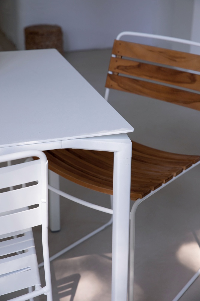 Detailaufnahme eines Fermob Surprising Teak Chair Outdoor Stuhls mit weißem Gestell und Teakholz-Sitzfläche neben einem weißen Gartentisch.