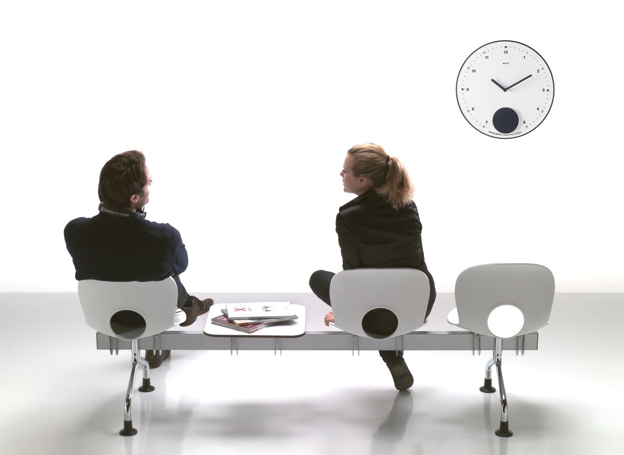Appuntamento Wanduhr von Rexite im Wartezimmer mit zwei Personen, die auf einer Bank sitzen.